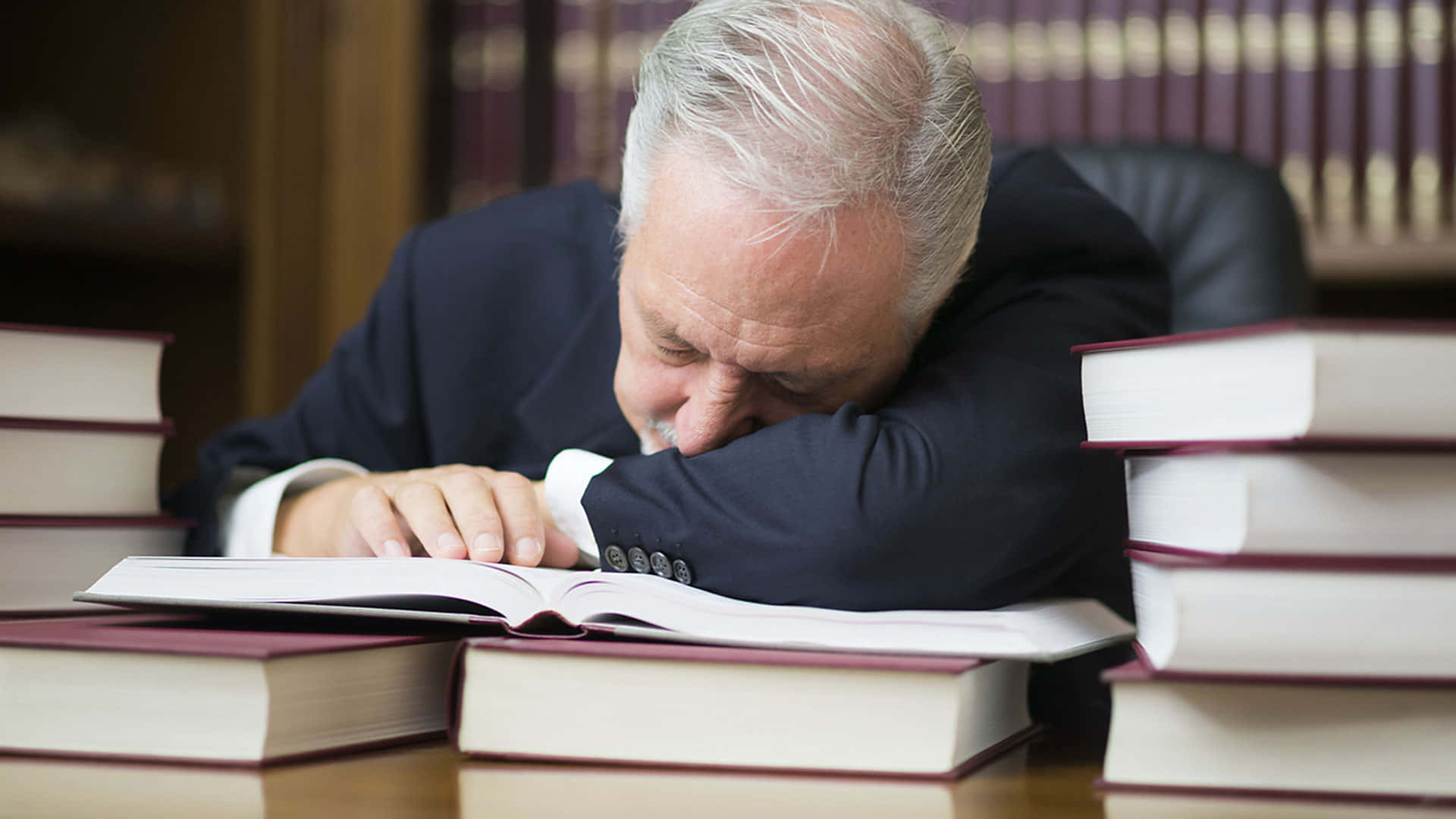 Sleeping 桌布 壁紙 — 一個穿著西裝的疲憊老人，將頭靠在一本打開的書上，周圍堆滿了高聳的法律書籍。