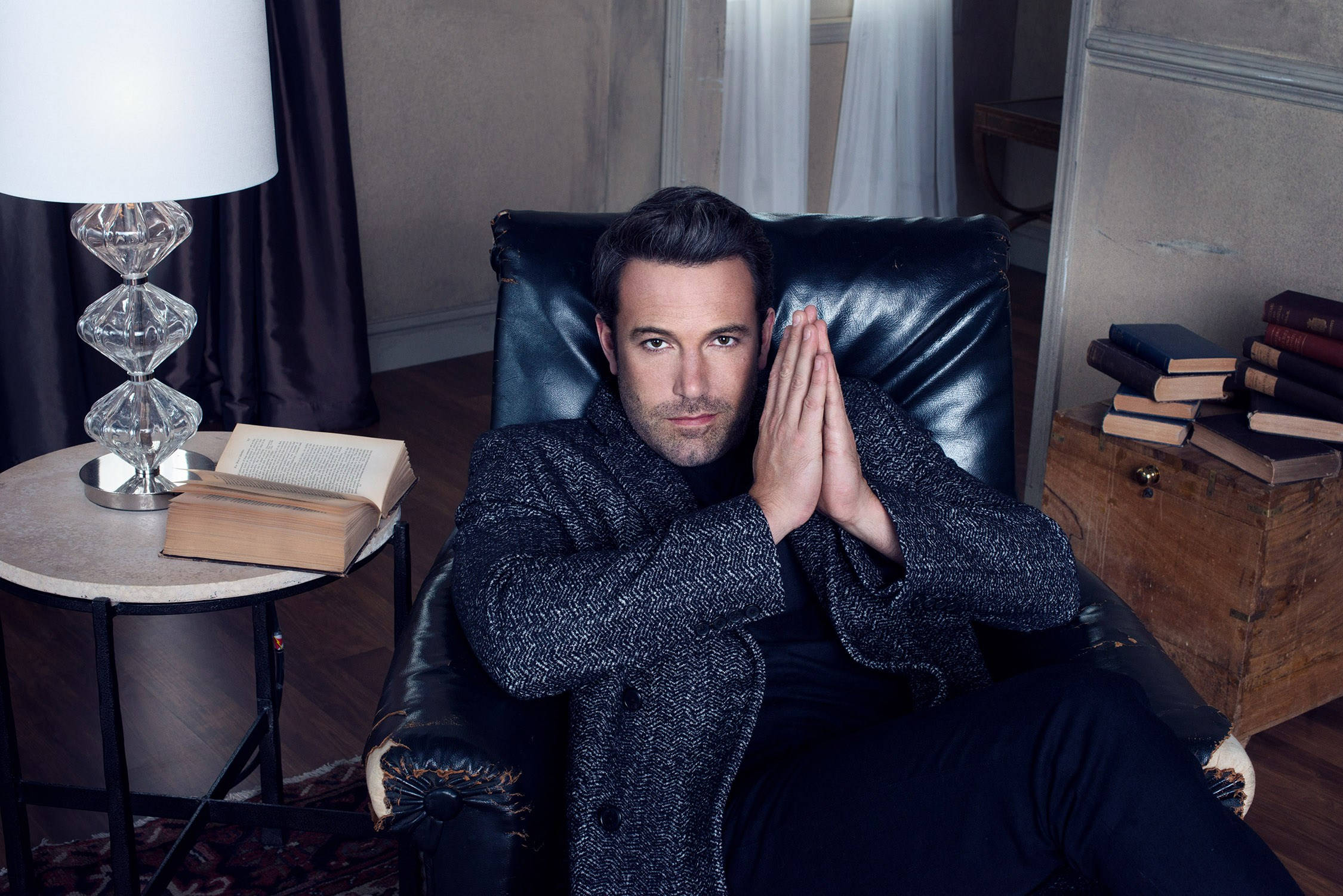Ben Affleck 桌布 壁紙 — 一個穿著質感外套的男人坐在黑色皮椅上，雙手緊握，周圍環繞著書籍和水晶燈，在一個舒適的房間裡。