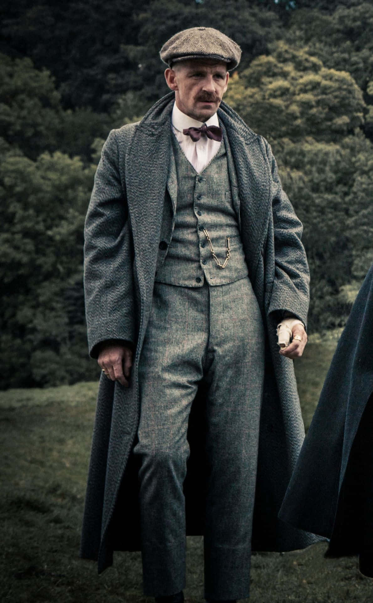 穿著復古灰色西裝和扁平帽的男人在森林中手持一把手槍。 — Peaky Blinders 壁紙