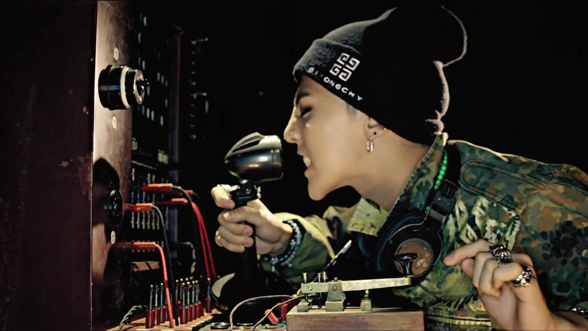 G-dragon 桌布 壁紙 — 一個穿著迷彩外套和毛帽的人，在復古收音機設備旁熱情地對著懷舊麥克風唱歌。