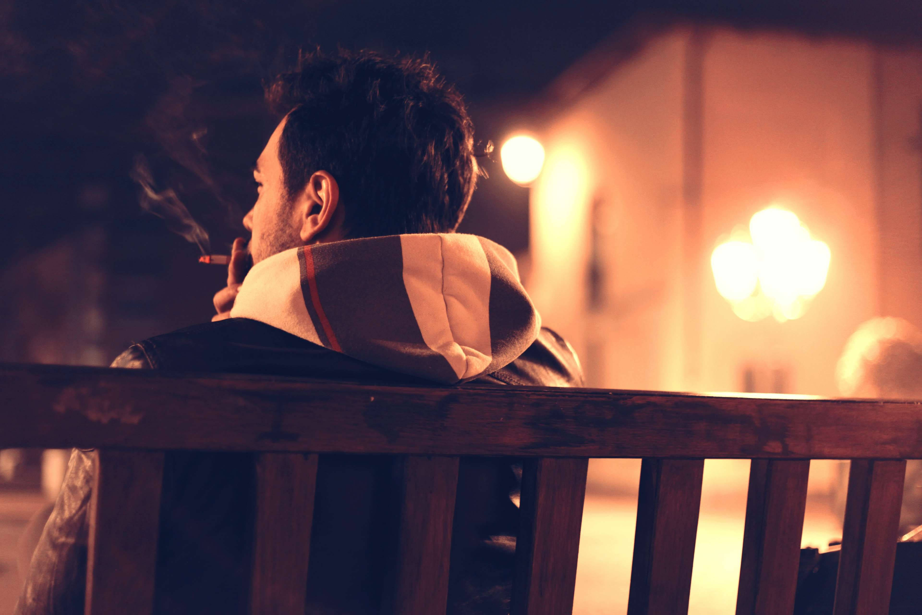 Alone 桌布 壁紙 — 一個穿著連帽外套的男人在木長椅上抽煙，背景是溫暖的夜燈和模糊的光暈。