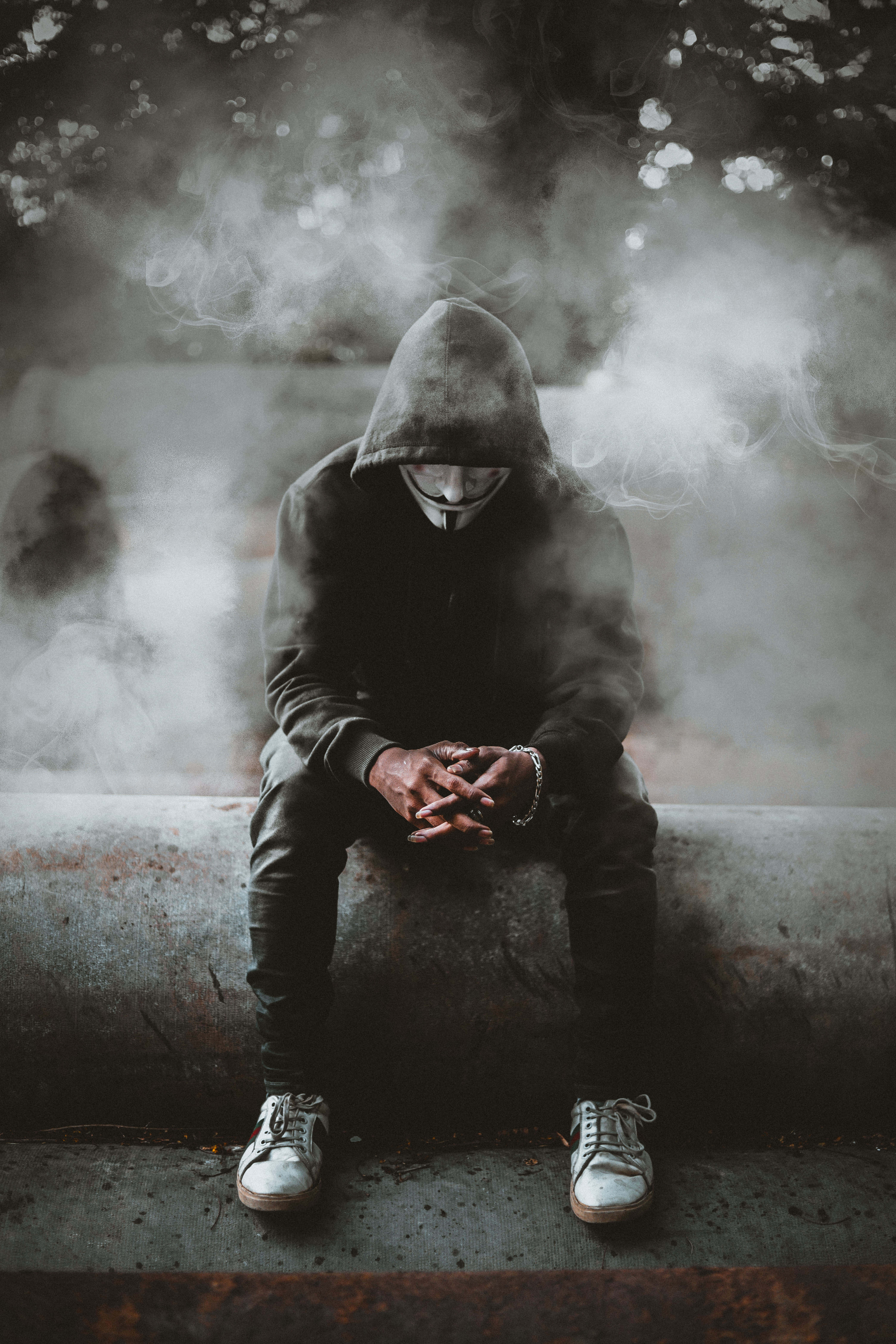Black Sad 桌布 壁紙 — 一個穿著連帽衫、戴著蓋伊·福克斯面具的人蜷縮在混凝土邊緣上，周圍是昏暗骯髒的城市環境中旋轉的煙霧。