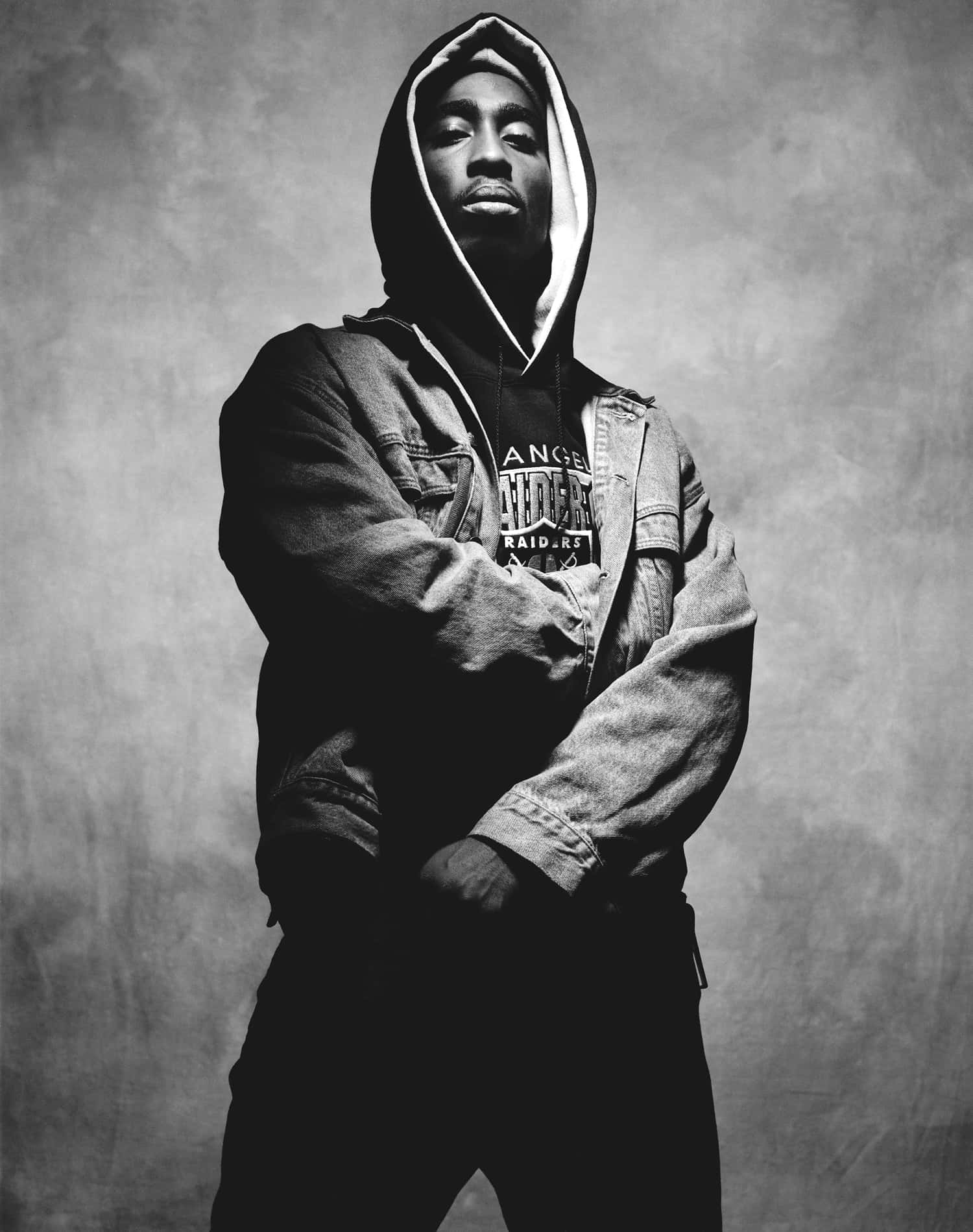 tupac 桌布 壁紙 — 一個穿著連帽衫和牛仔外套的男人，雙手交叉，站在有紋理的背景前的黑白肖像。