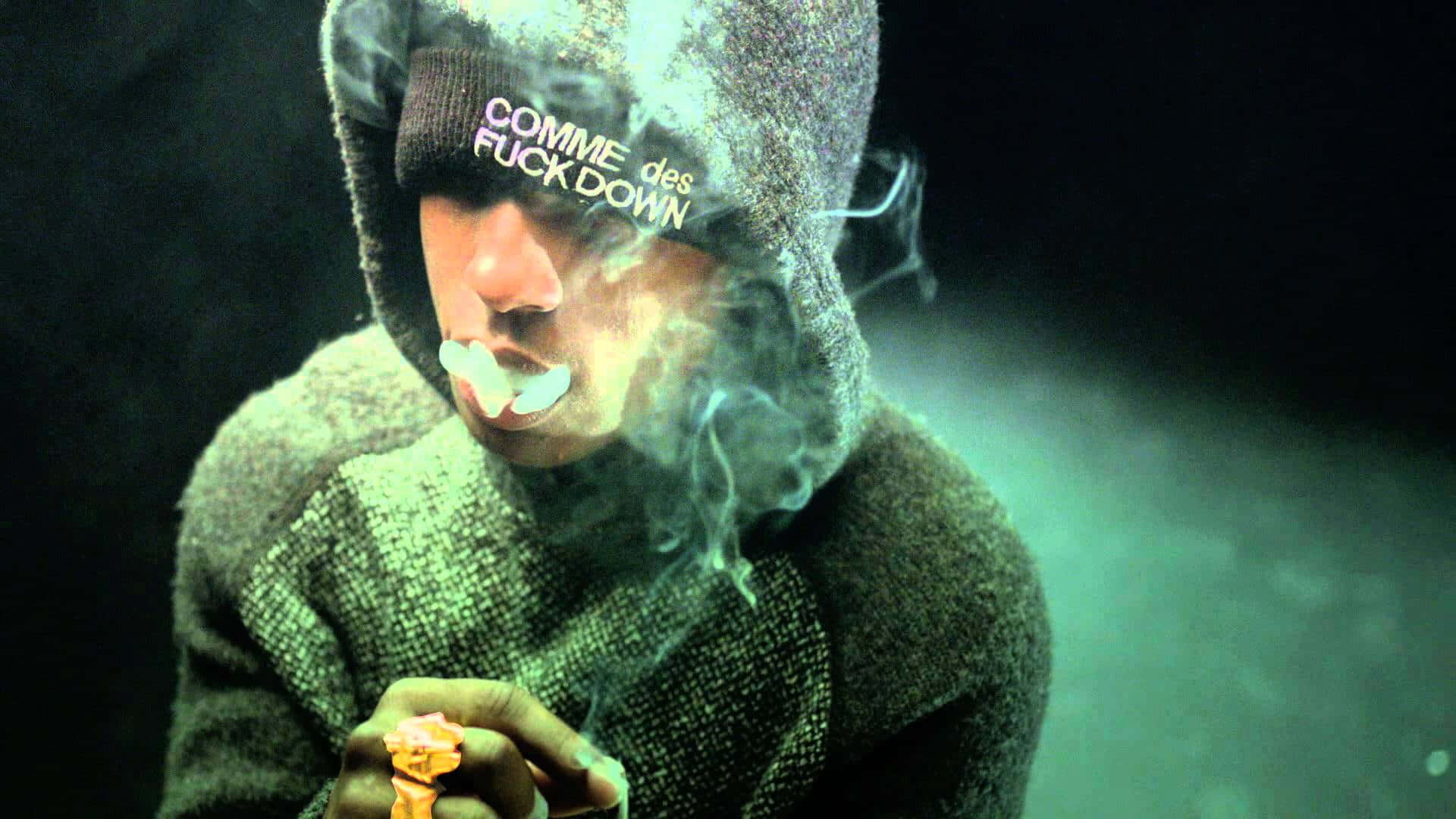 Asap Rocky 桌布 壁紙 — 一個穿著連帽衫的人，在深綠色朦朧的背景前，手持點燃的大麻煙並呼出煙霧。