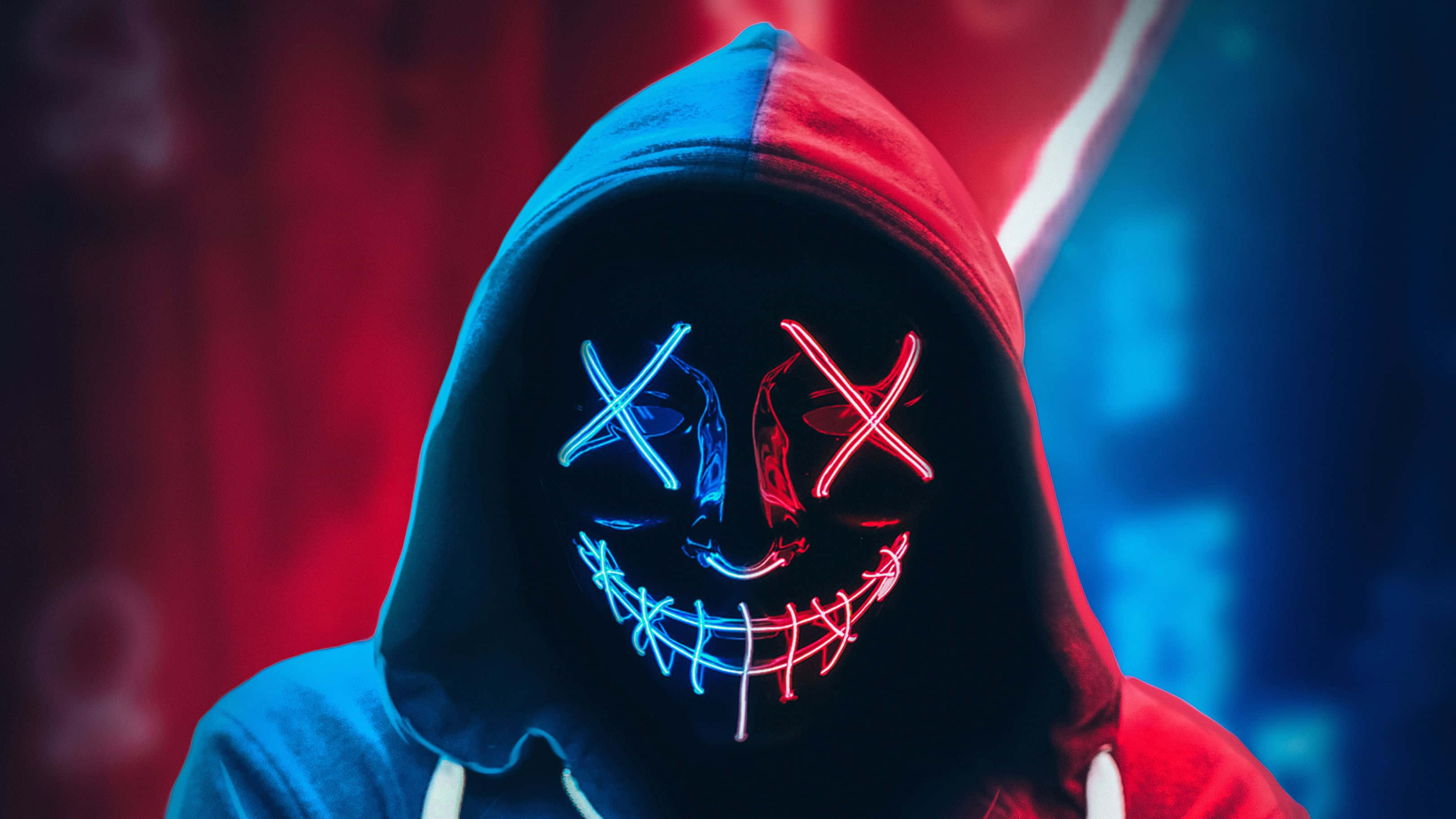 Mask 桌布 壁紙 — 一個穿著連帽衫的人戴著發光的霓虹面具，面具上有X形眼睛和縫合的笑容，背景是紅色和藍色的模糊效果。