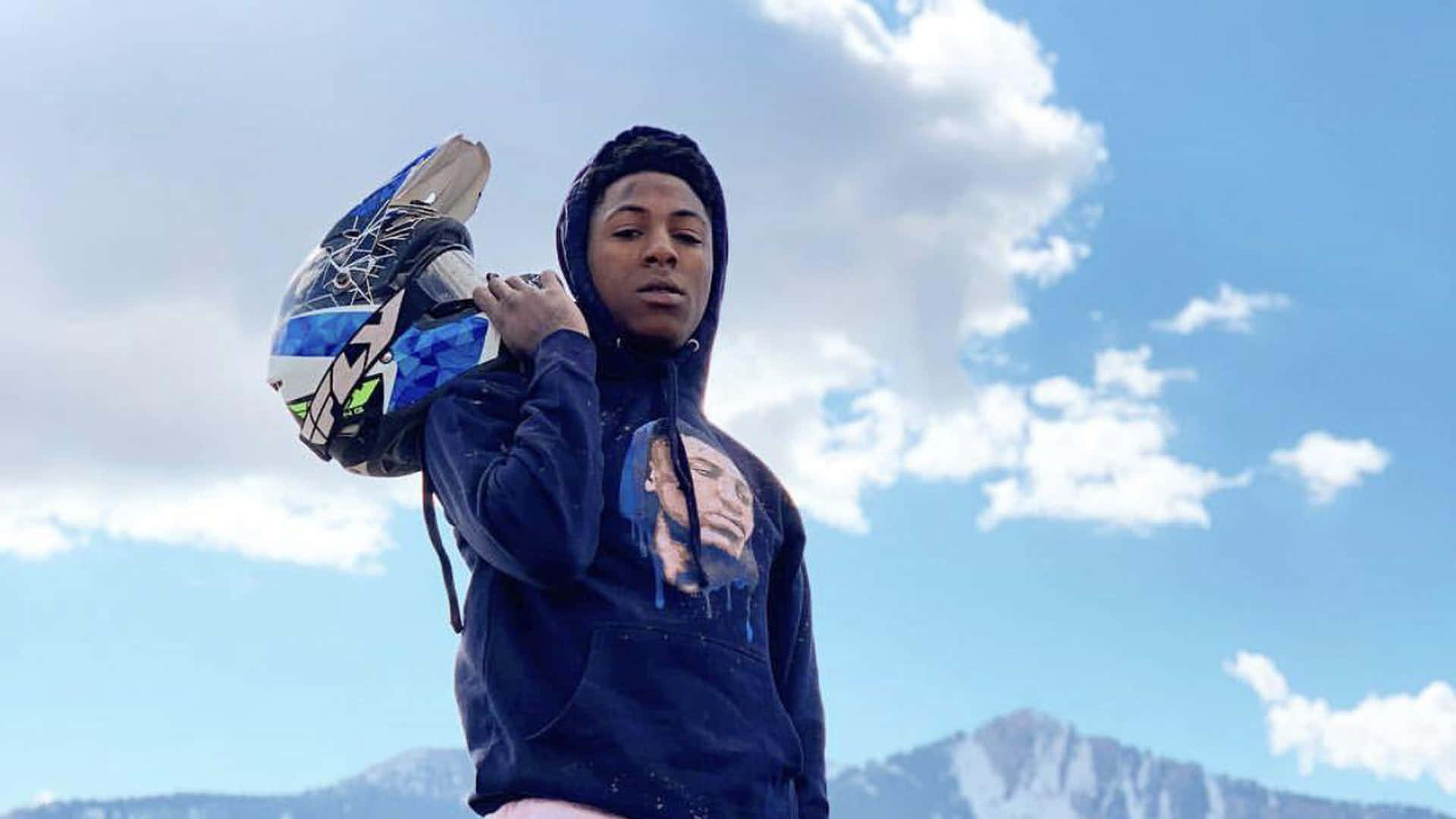 NBA YoungBoy 桌布 壁紙 — 一個穿著連帽衫的年輕人，手持滑雪板頭盔，背景是白雪覆蓋的山脈和藍天白雲。