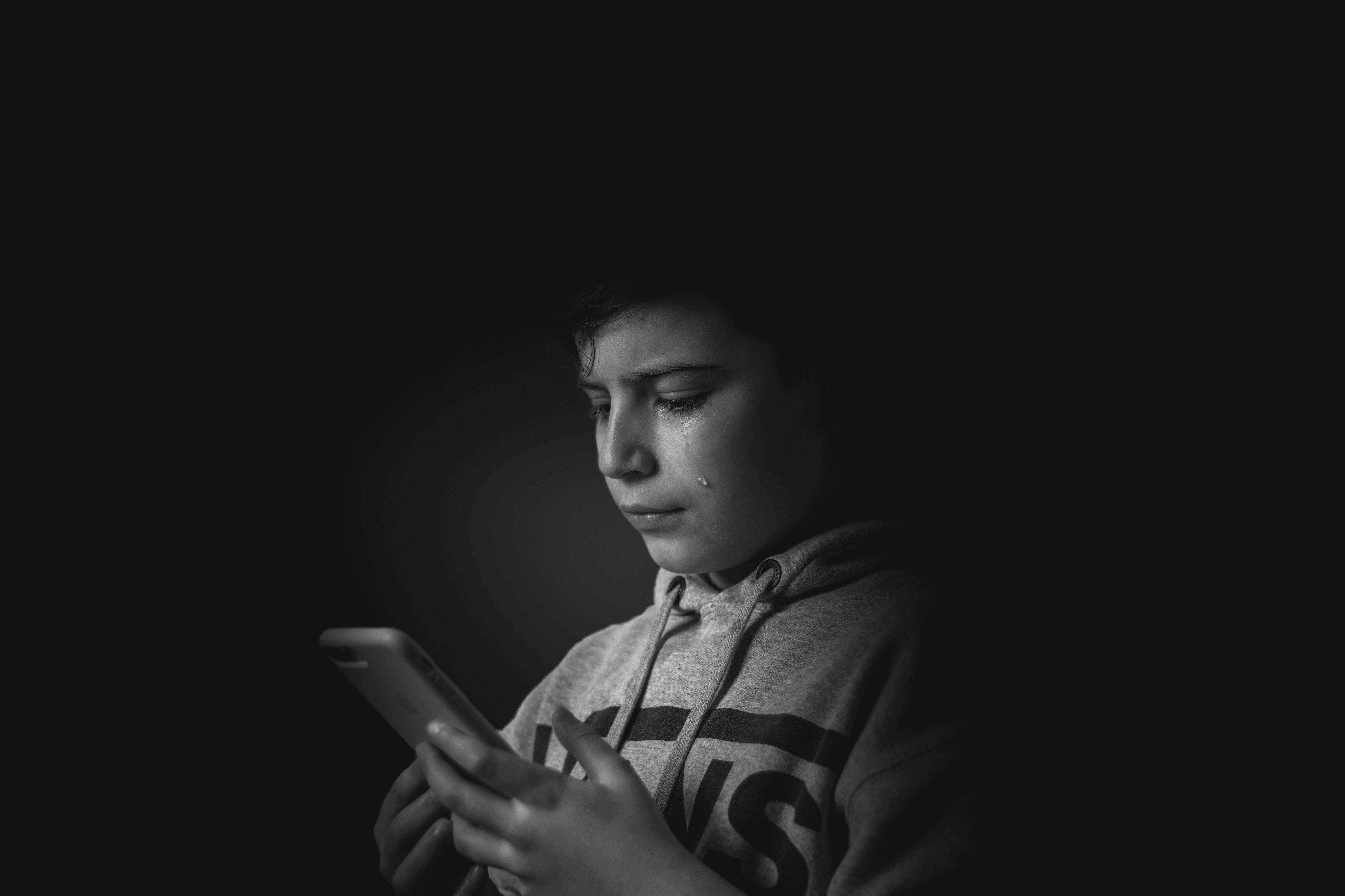 crying sad boy 桌布 壁紙 — 一個穿著連帽衫的年輕男孩在黑暗背景下盯著手機，淚水順著臉頰滑落。