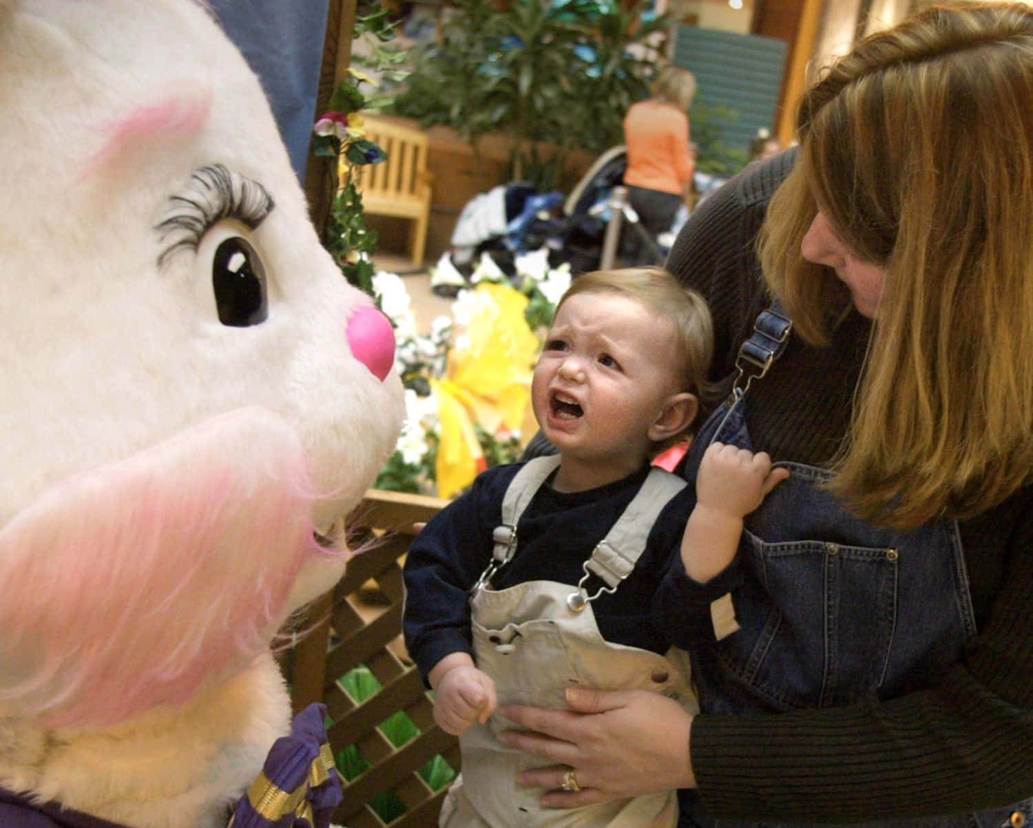 Creepy Easter Bunny 桌布 壁紙 — 一個穿著連身衣的幼兒被一個女人抱著，面對一個大粉紅耳朵復活節兔子吉祥物時哭泣。