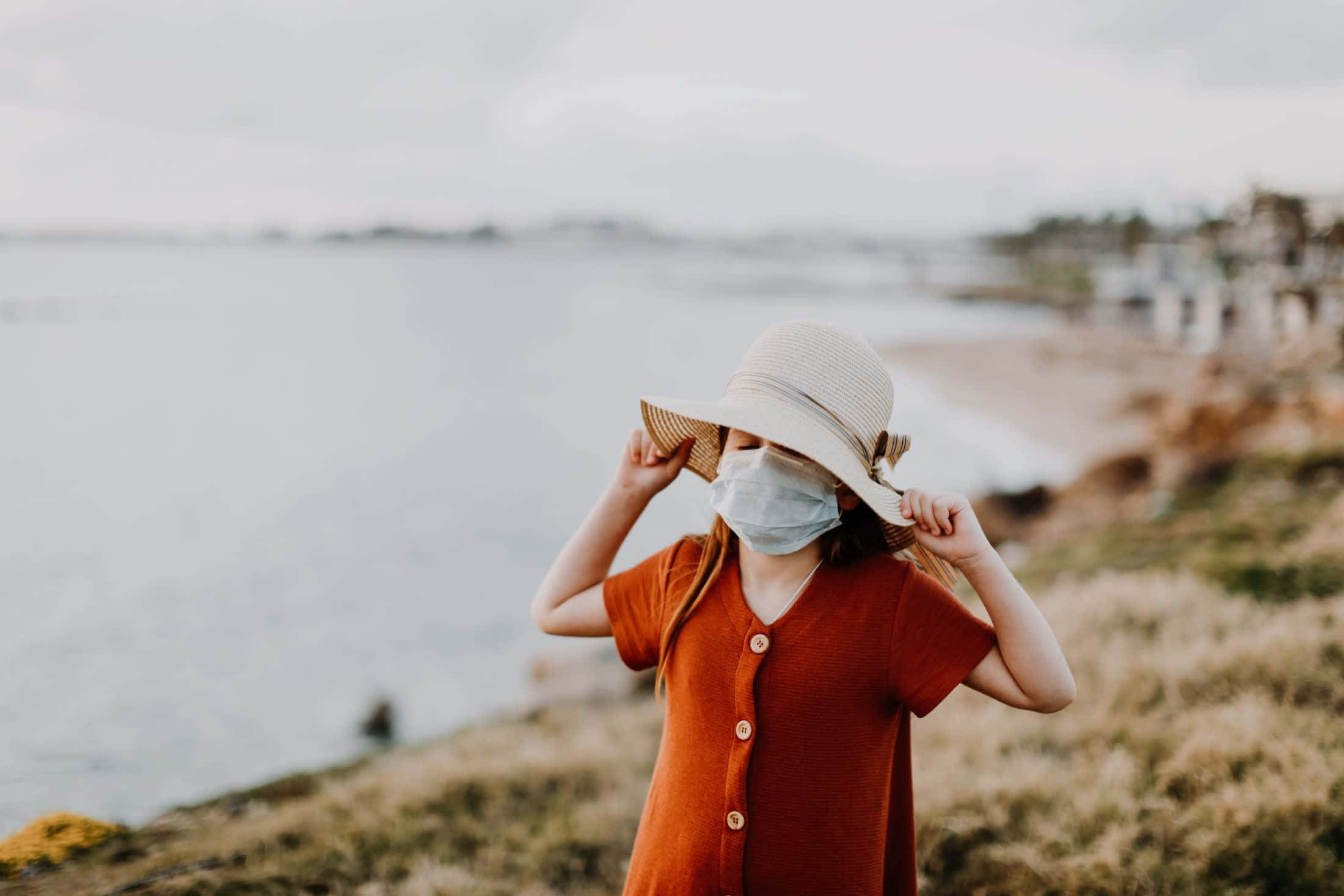 Scenery 桌布 壁紙 — 一個穿著鏽色連衣裙和寬邊帽的小女孩，站在一個俯瞰平靜大海的草崖上，在陰天中調整她的口罩。