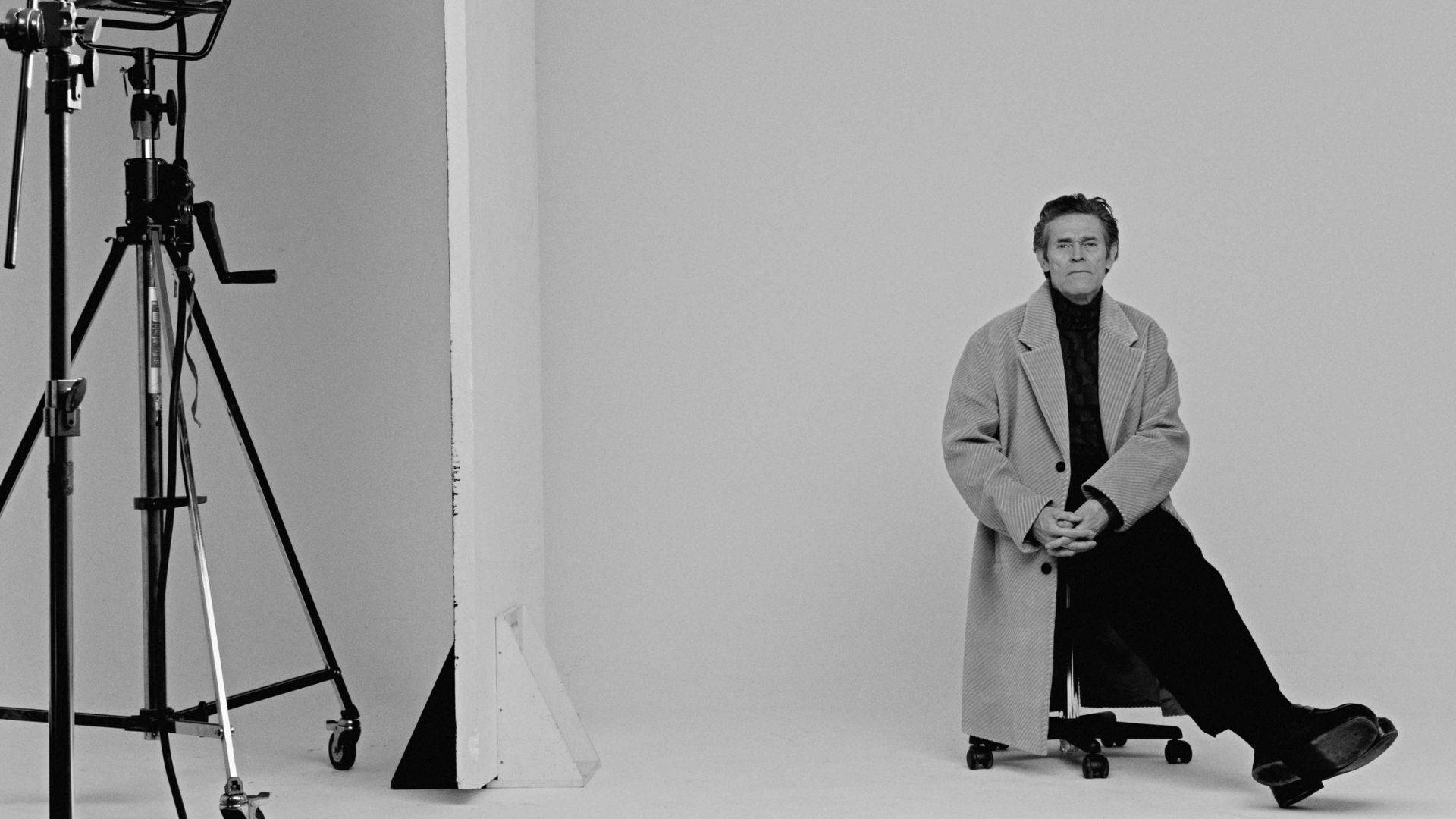 Willem Dafoe 桌布 壁紙 — 一個穿著長外套的男人坐在滾動椅上，身處一個簡約的工作室，可見照明設備。