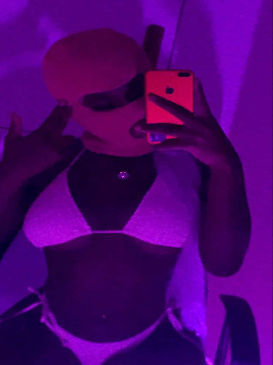 Ski Mask Girl 桌布 壁紙 — 一個穿著閃亮比基尼的人在強烈紫色燈光下，用發光紅色手機拍鏡子自拍照。