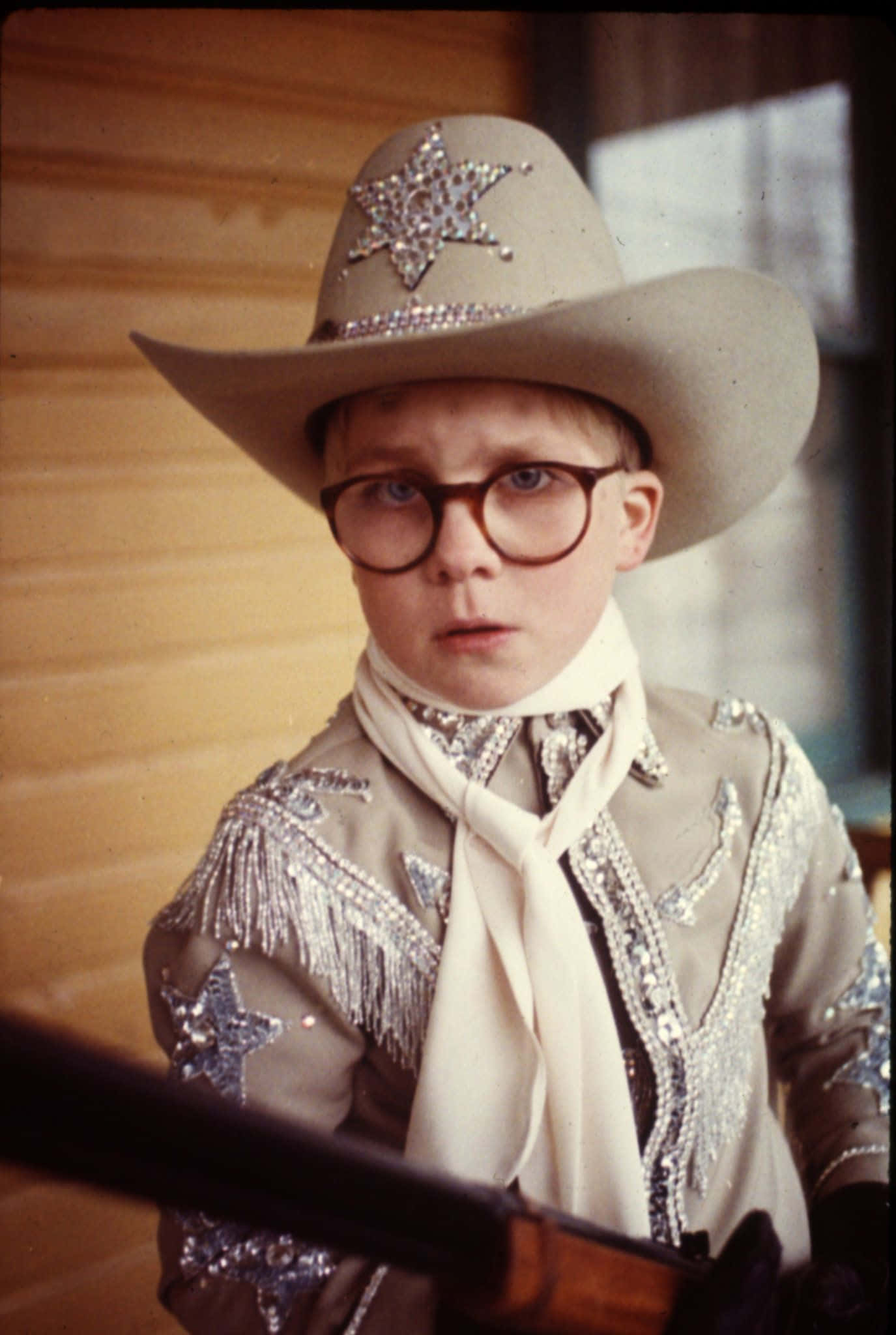 a christmas story 桌布 壁紙 — 一個穿著閃亮牛仔裝束和眼鏡的小男孩，在室內專注地盯著鏡頭，手持步槍。