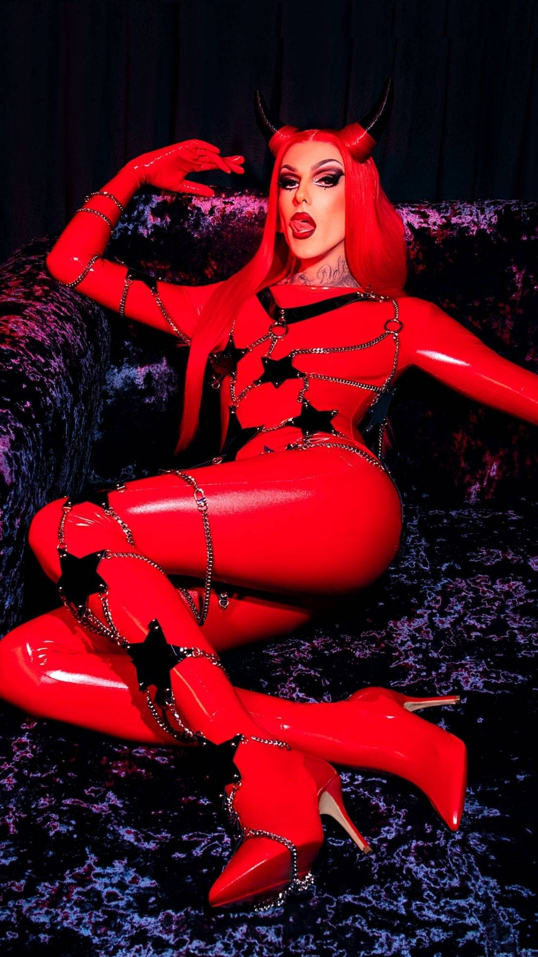 Jeffree Star 桌布 壁紙 — 一個穿著閃亮紅色魔鬼服裝、頭上有角的人，在戲劇性燈光下挑釁地躺在天鵝絨沙發上。
