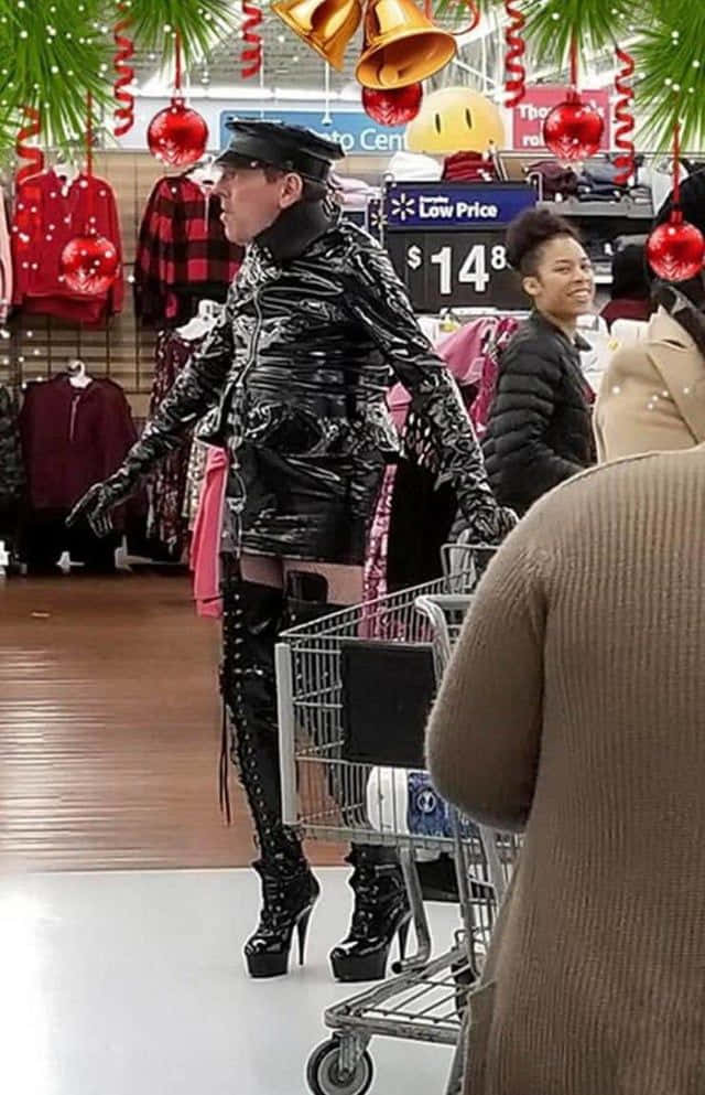 meme 桌布 壁紙 — 一個穿著閃亮黑色皮革服裝和高跟鞋的人推著購物車穿過節日假期商店的通道。
