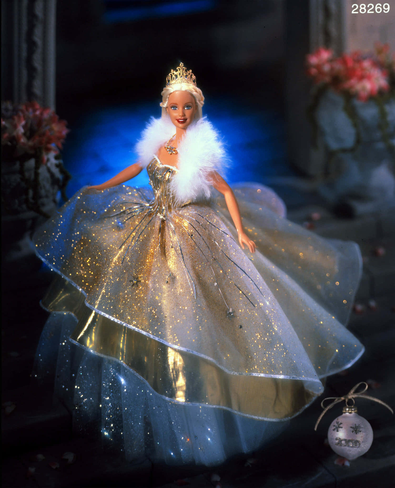 Barbie Doll 桌布 壁紙 — 一個穿著閃閃發光的金色和藍色禮服、白色毛領和皇冠的芭比娃娃，在深藍色背景和花卉佈置前優雅地站立。