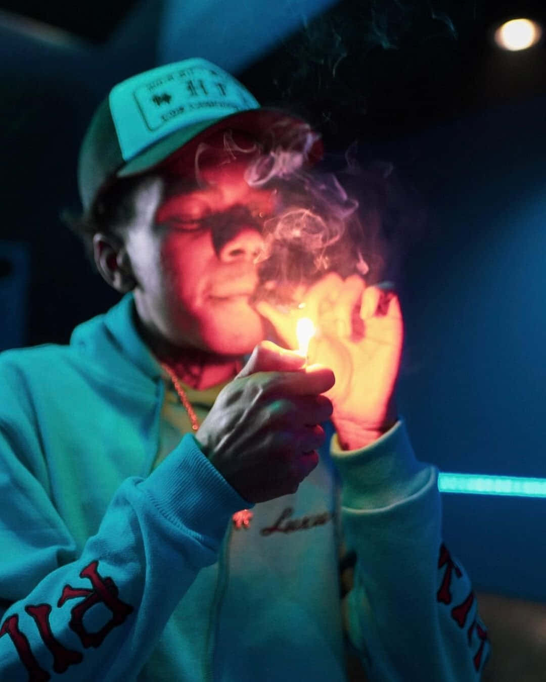 說唱歌手 桌布 壁紙 — 一個穿著青綠色連帽衫和帽子的人，在一個光線昏暗、帶有藍色霓虹燈光和煙霧繚繞的房間裡點燃一支香煙。