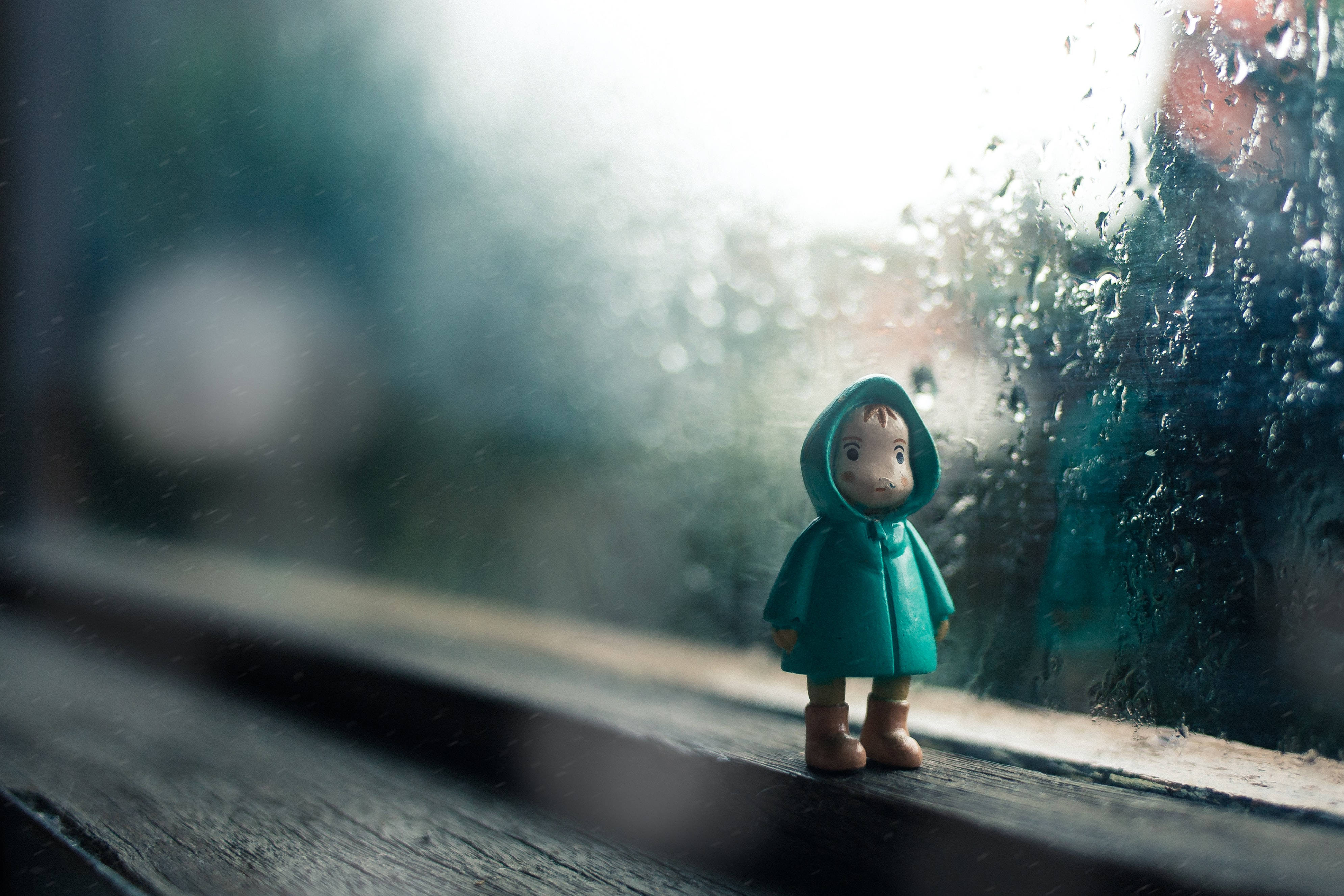 動態桌面 桌布 壁紙 — 一個穿著青綠色雨衣的小人偶獨自站在窗台上，凝視著窗外雨濛濛、模糊的世界。