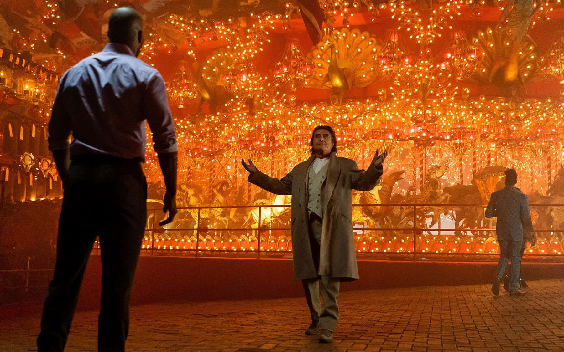 American Gods 桌布 壁紙 — 一個穿著風衣的男人在夜晚張開雙臂站在燈光璀璨的旋轉木馬前，面對另一個穿著藍色襯衫的男人。