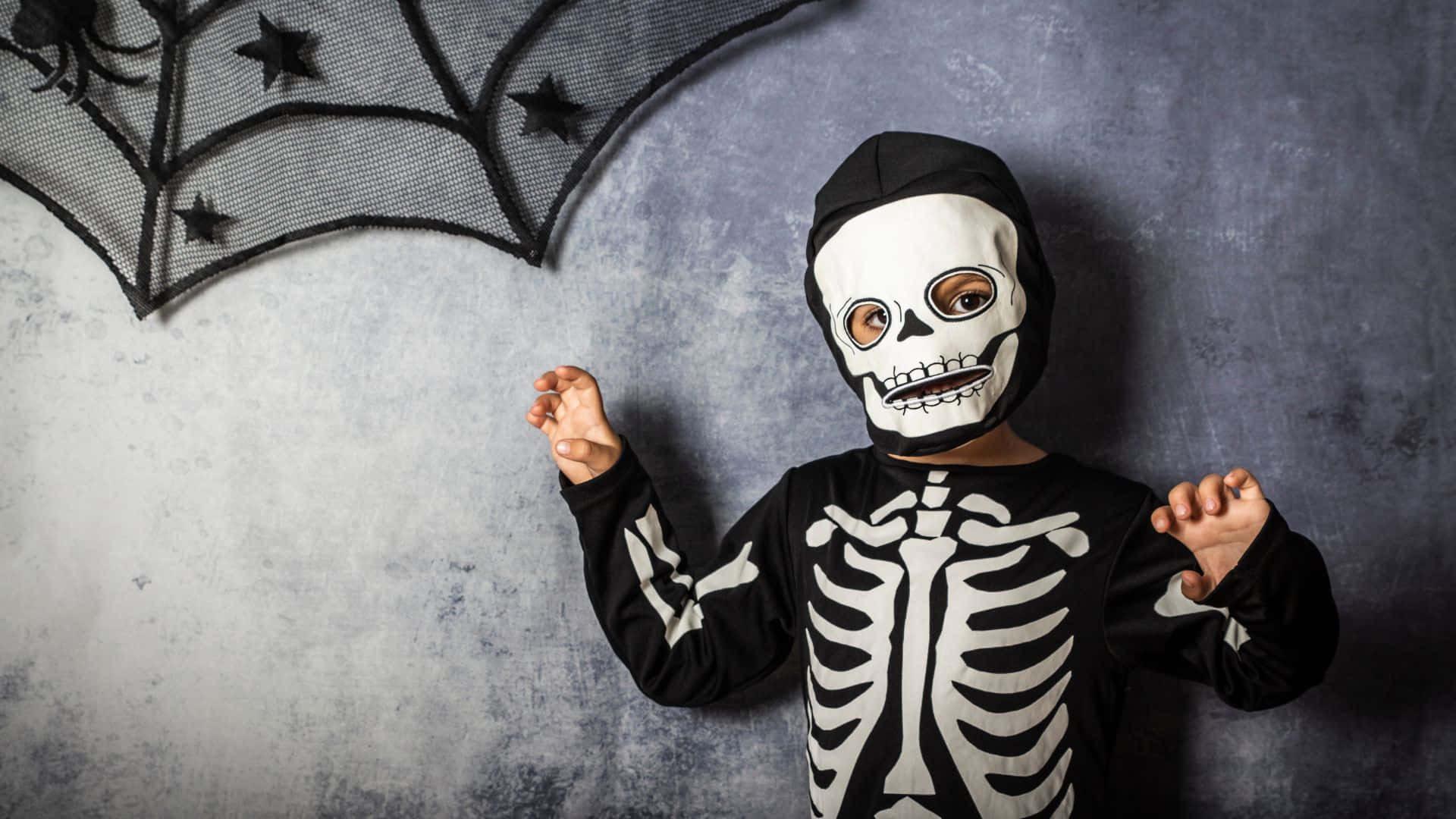 Skeleton Costumes 桌布 壁紙 — 一個穿著骷髏服裝、戴著骷髏面具的孩子站在灰色牆壁前，上方有黑色蜘蛛網裝飾。