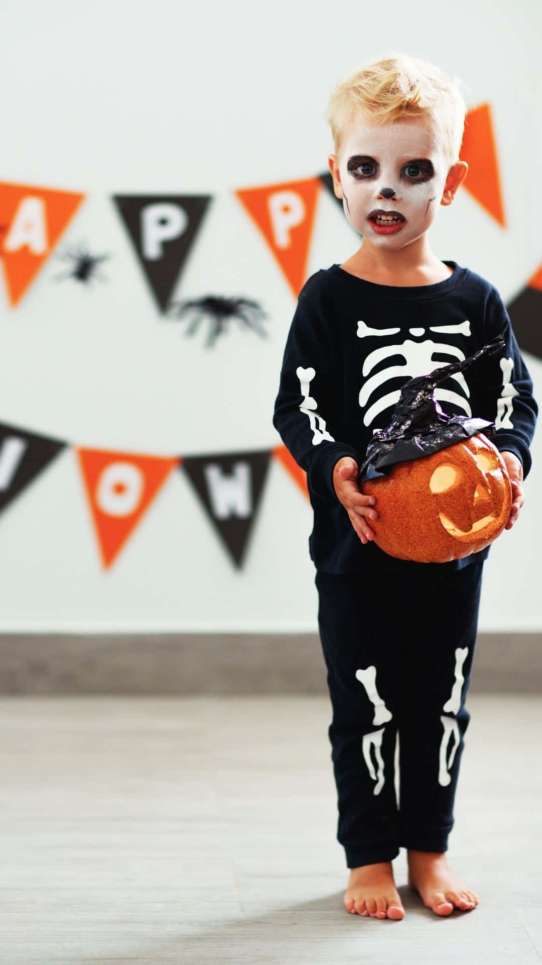 Skeleton Costumes 桌布 壁紙 — 一個穿著骷髏服裝的小孩在「HAPPY HALLOWEEN」橫幅下拿著發光的南瓜燈。