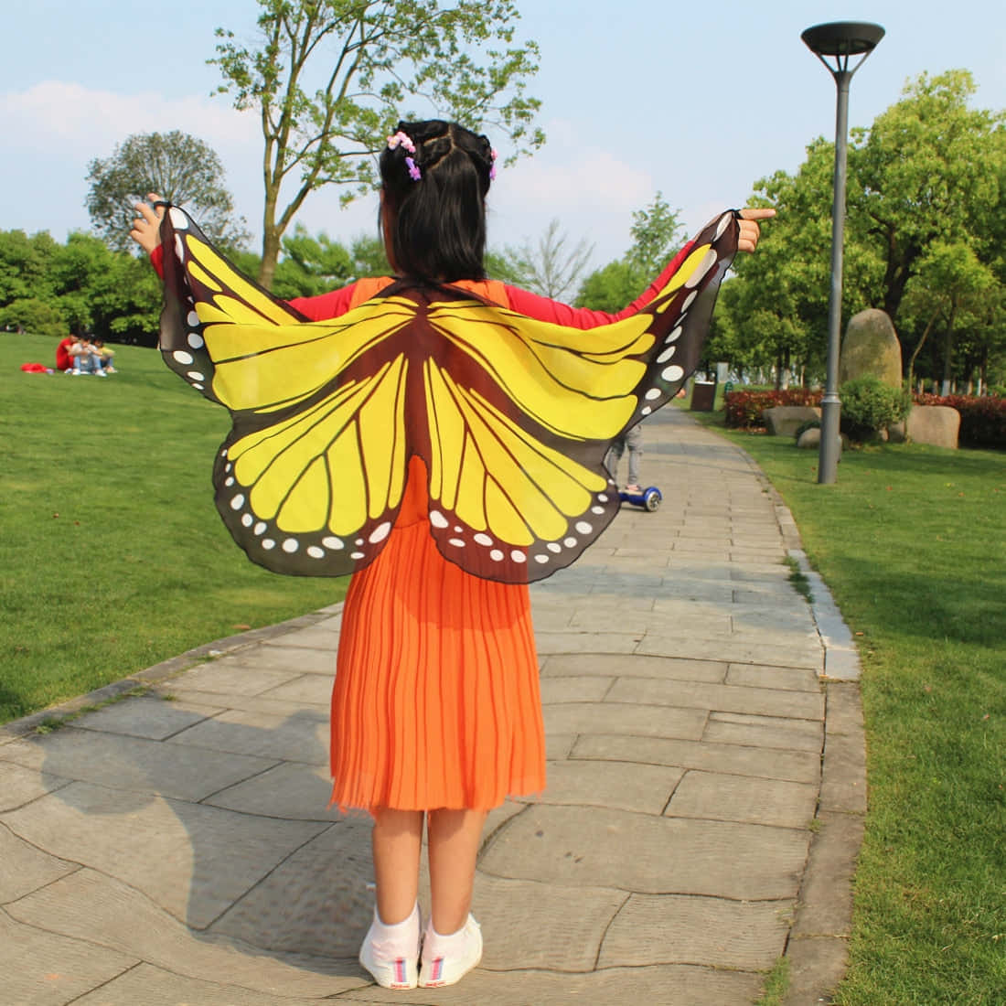 Butterfly Wing Dress 桌布 壁紙 — 一個穿著鮮橙色連衣裙的孩子站在公園小徑上，戴著大大的黃黑蝴蝶翅膀。