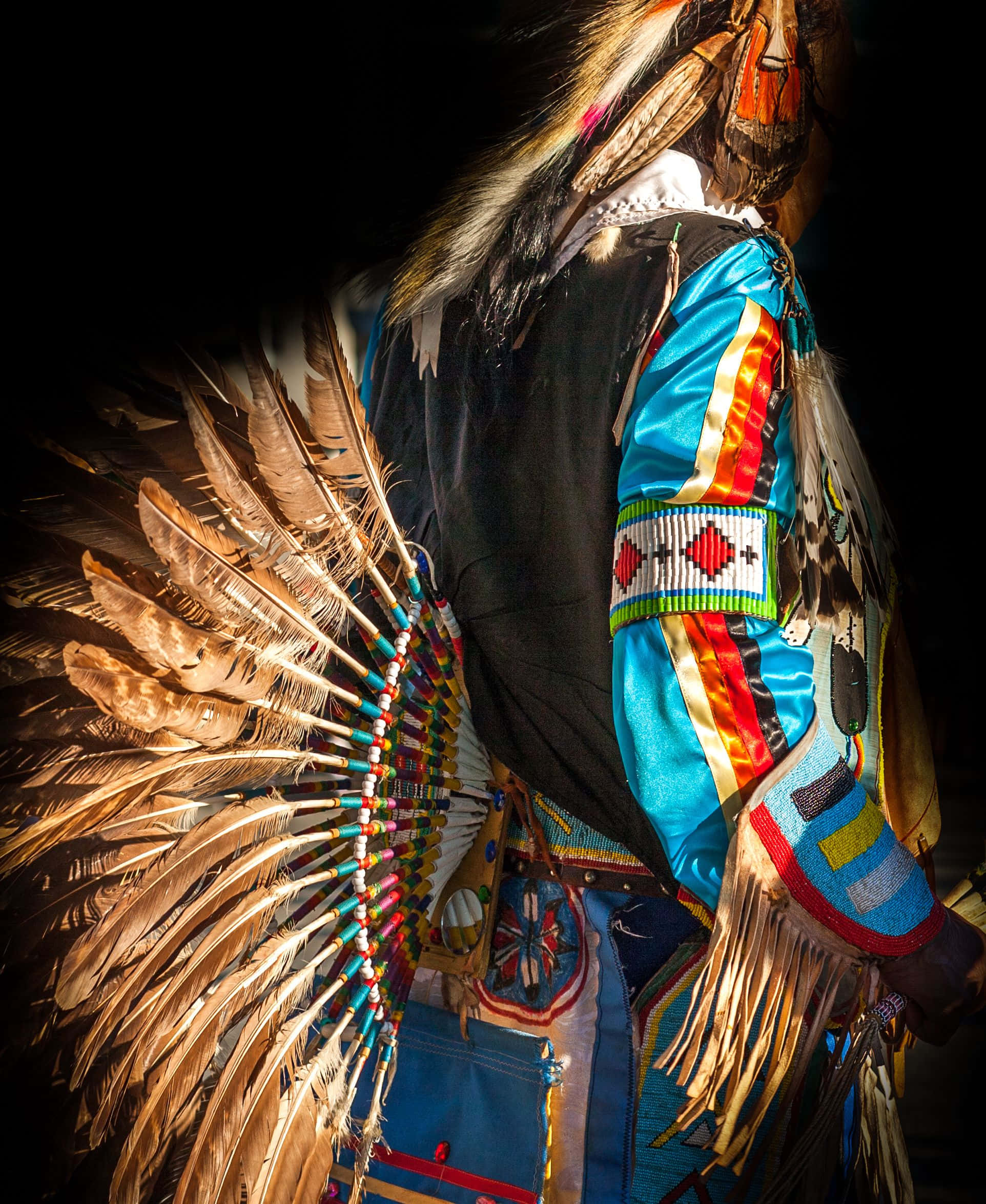 Native American 桌布 壁紙 — 一個穿著鮮豔傳統美洲原住民服飾、戴著大型羽毛頭飾和珠飾圖案的人，在黑色背景的戲劇性聚光燈下站立。