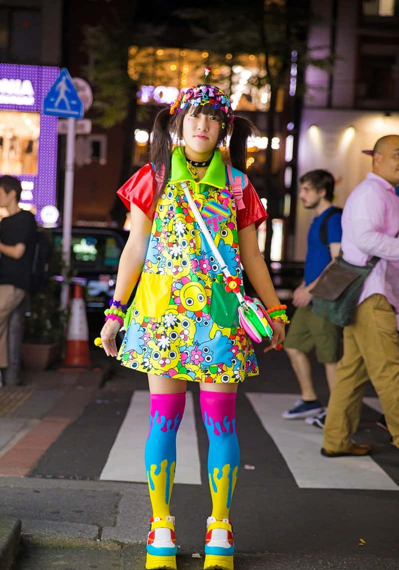 扎染 桌布 壁紙 — 一個穿著鮮豔多彩服裝、帶有卡通圖案和明亮腿部彩繪的人，在夜晚的城市街道上自信地站立。