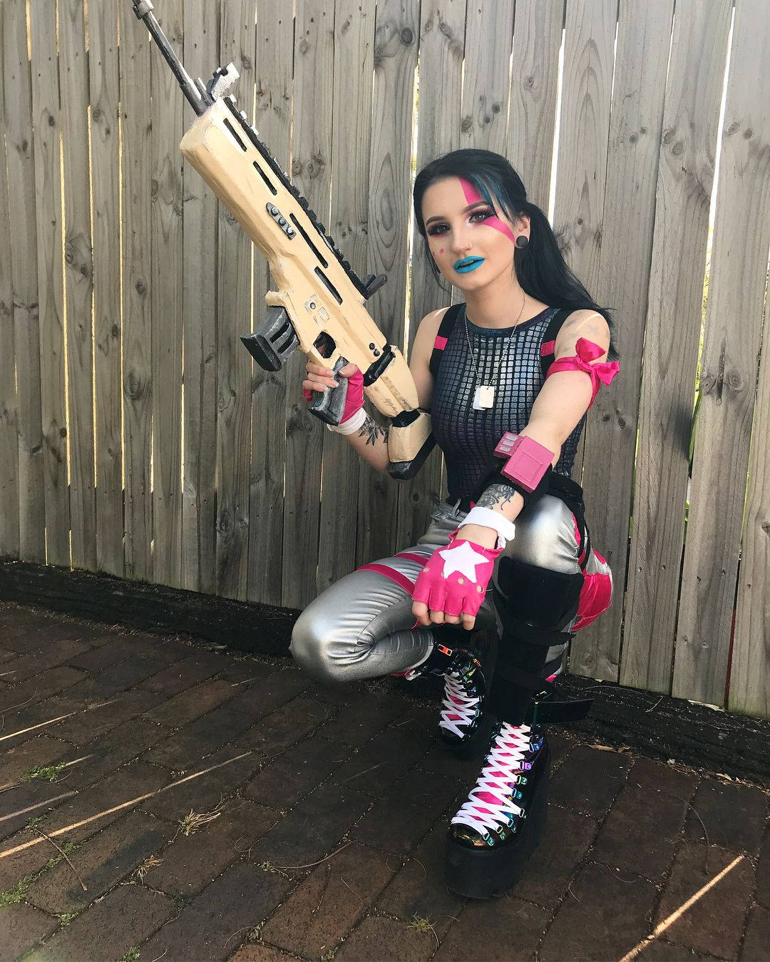 Sparkle Specialist Fortnite 桌布 壁紙 — 一個穿著鮮豔粉紅和銀色哈莉·奎茵cosplay的人蹲著，手持大型玩具槍靠著木柵欄。