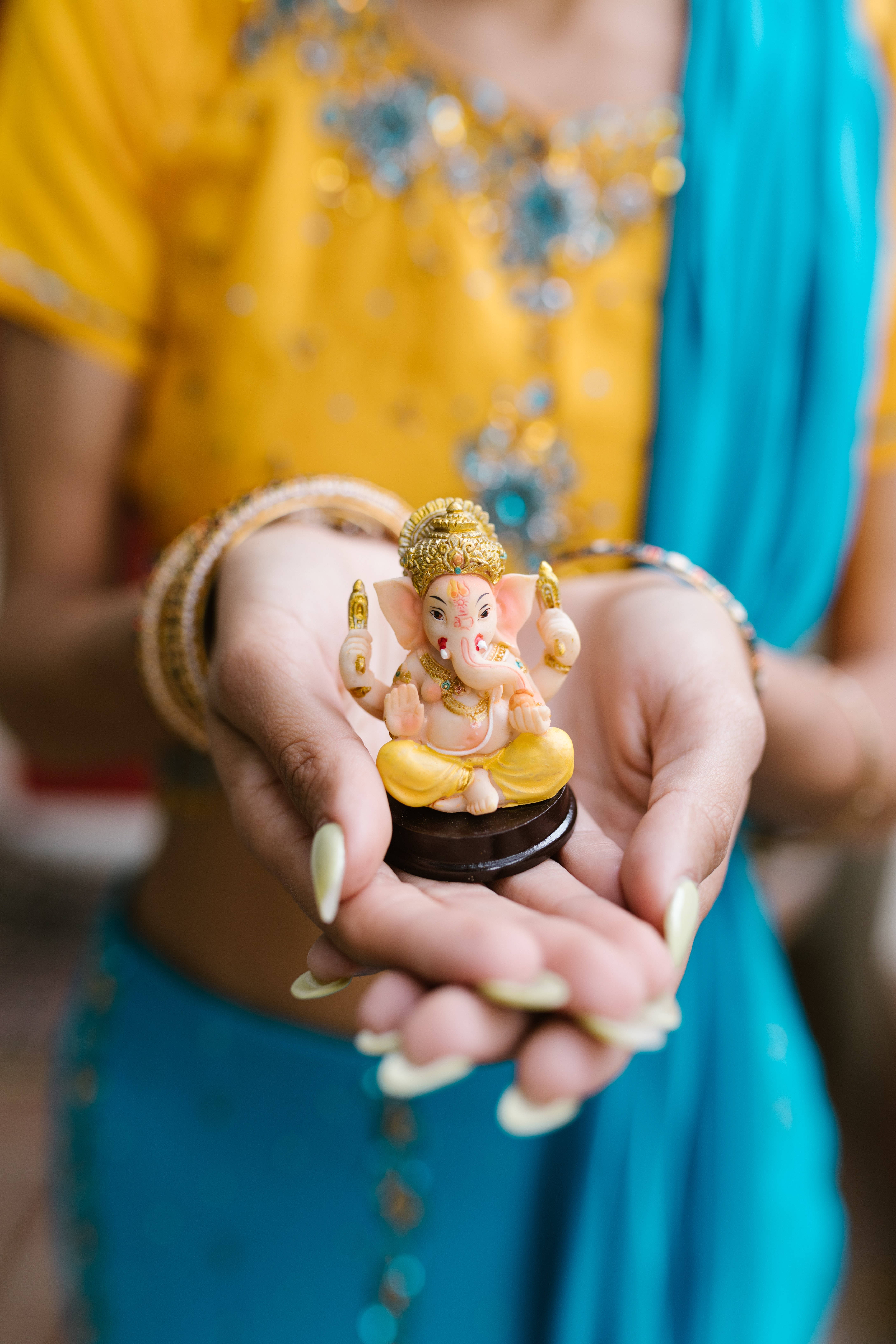 ganesh iPhone 桌布 壁紙 — 一個穿著鮮豔黃色和藍色傳統服飾的人，雙手輕輕捧著一個小小的金色象神伽內什雕像。