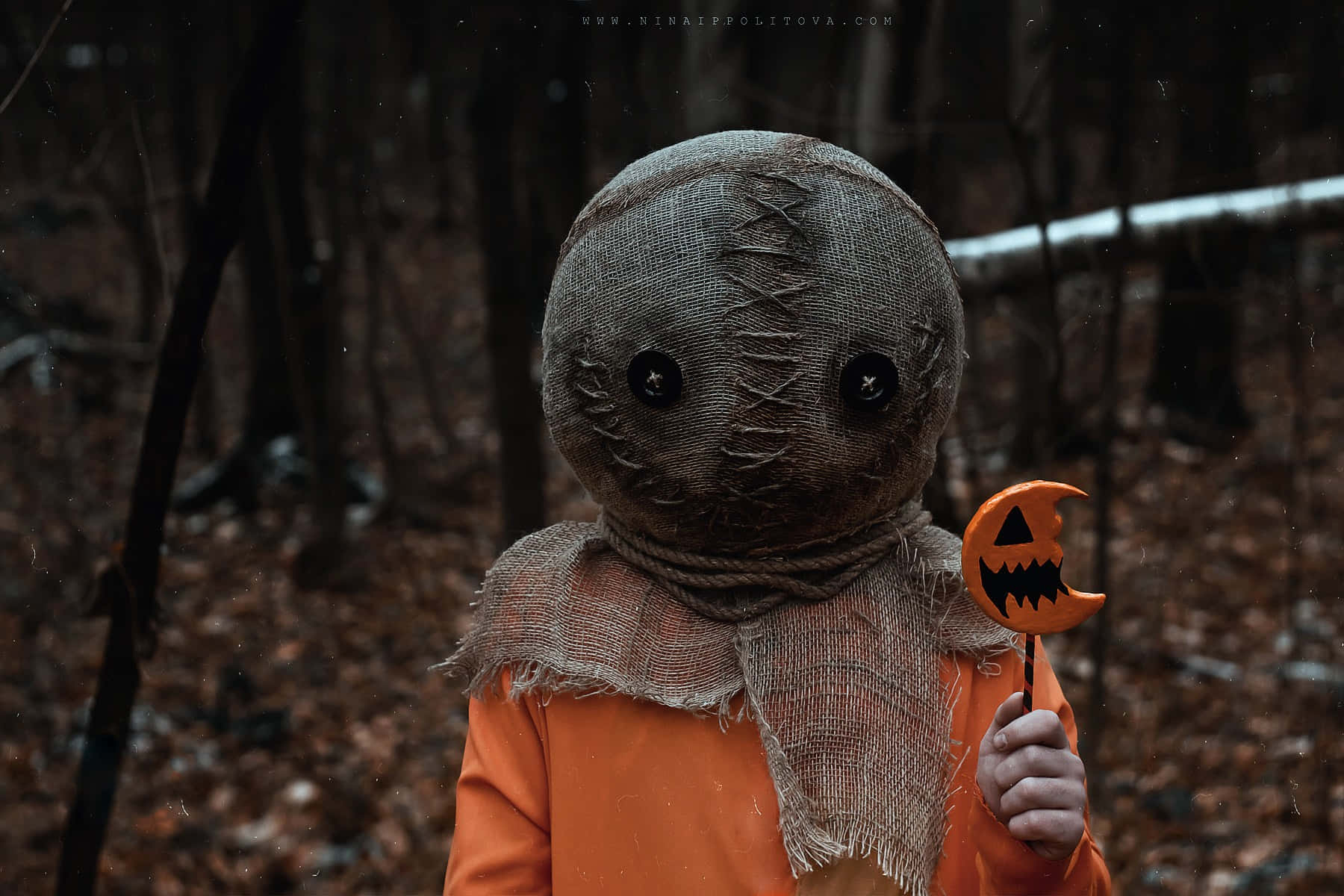 Trick Or Treat 桌布 壁紙 — 一個穿著麻布袋服裝、有黑色鈕扣眼睛的小孩，在雪地森林中拿著一個南瓜燈棒棒糖。