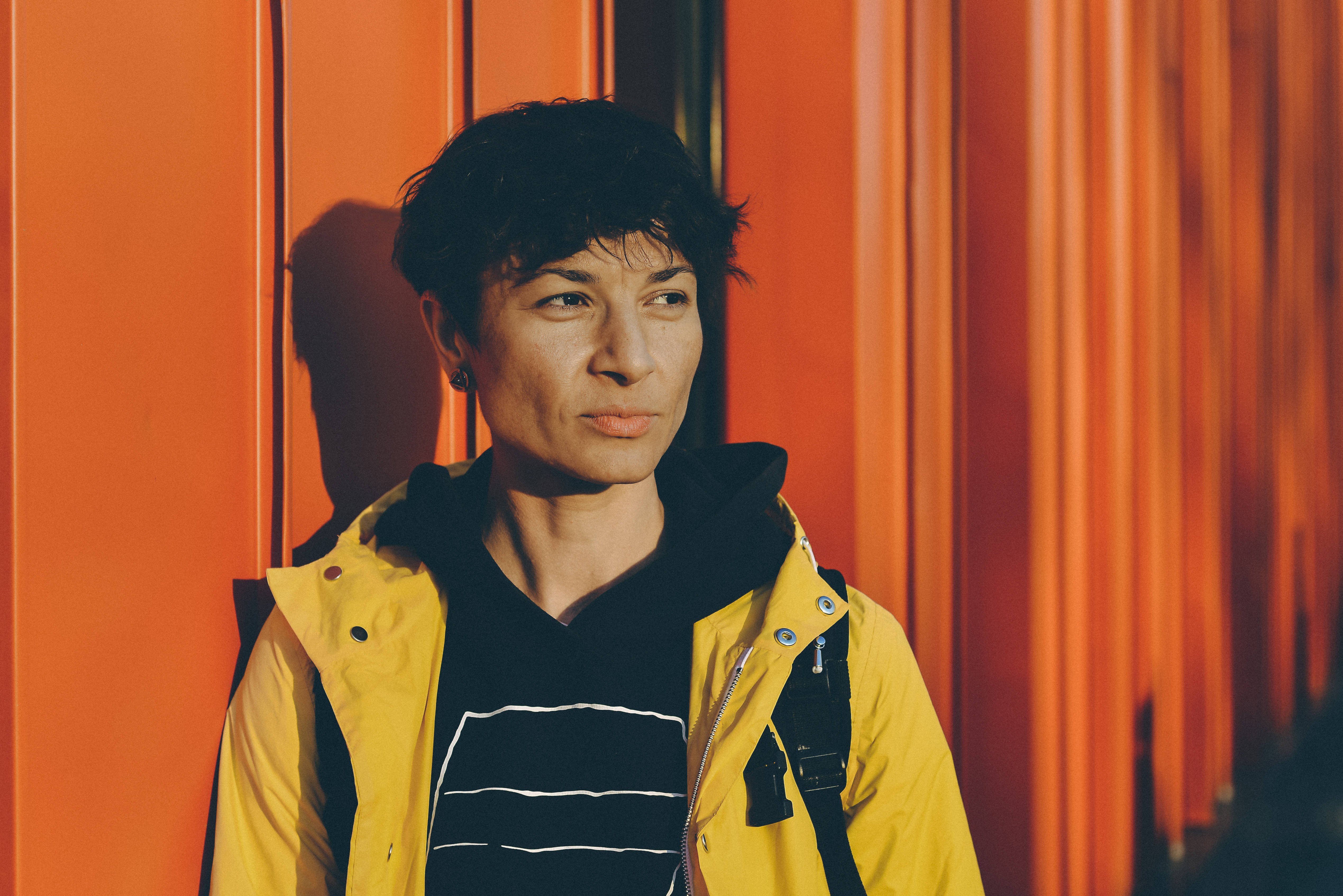 Tomboy 桌布 壁紙 — 一個穿著黃色外套的人站在充滿活力的橙色波紋牆前，沐浴在溫暖的陽光下。