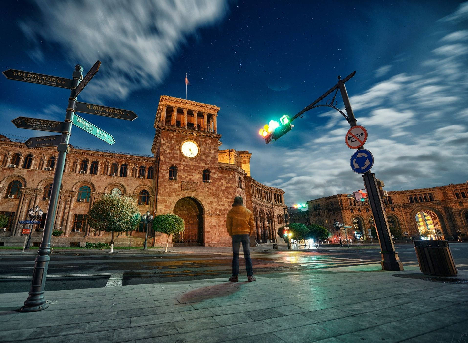 Armenia 桌布 壁紙 — 一個穿著黃色外套的孤獨身影，在星光燦爛的夜晚十字路口，站在歷史悠久的鐘樓下，交通燈閃爍著光芒。