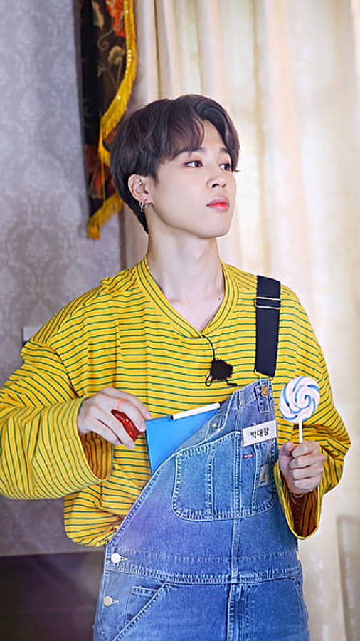 BTS 桌布 壁紙 — 一個穿著黃色條紋襯衫和牛仔工裝褲的年輕男子，手持棒棒糖和藍色筆記本，向上看。