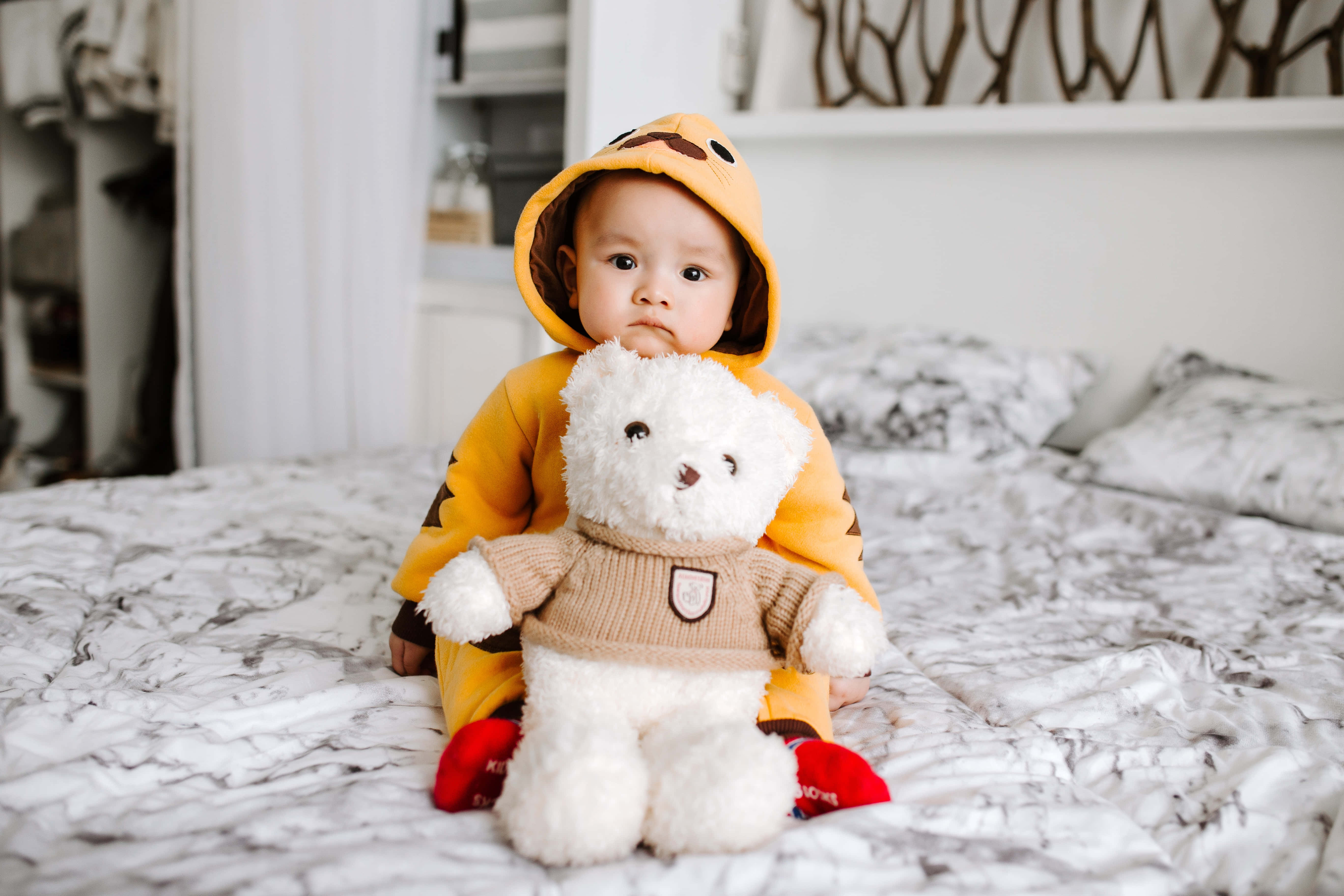 Baby Boy 桌布 壁紙 — 一個穿著黃色老虎連體衣的嬰兒坐在大理石花紋床上，抱著一隻穿著米色毛衣的白色泰迪熊。