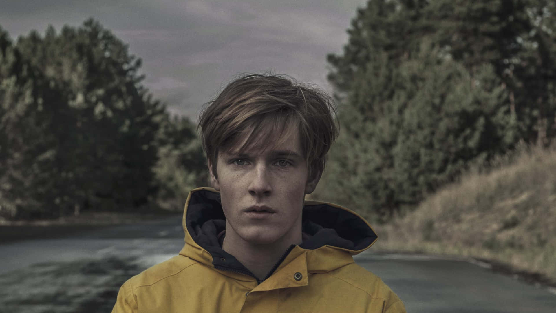 Dark Netflix 桌布 壁紙 — 一個穿著黃色連帽外套的年輕男子，在陰天之下，莊嚴地站在有松樹的河邊。