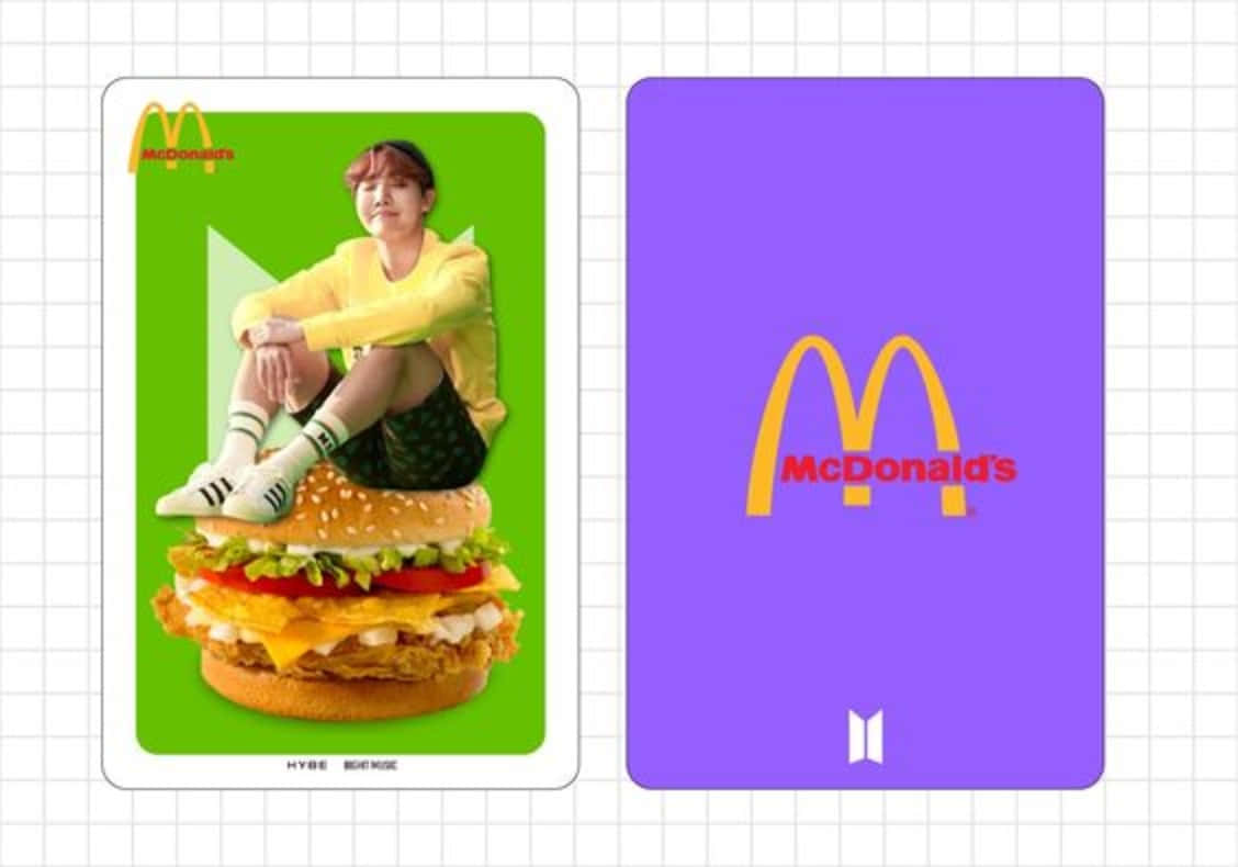 Bts Mac 桌布 壁紙 — 一個穿著黃色連帽衫的人坐在綠色卡片上的一個巨型漢堡上，旁邊是一張帶有BTS標誌的紫色麥當勞卡片。