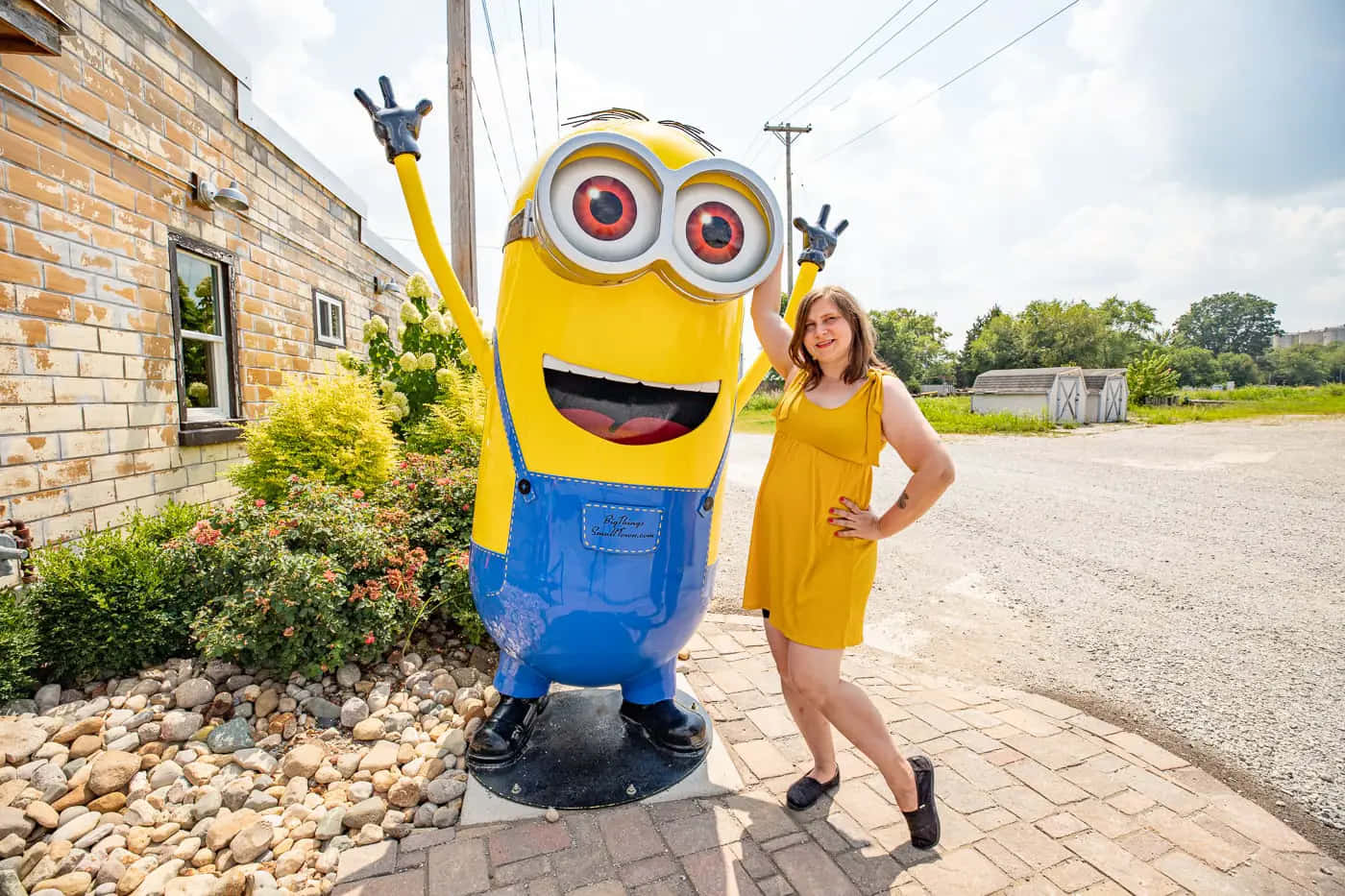 Minion 桌布 壁紙 — 一個穿著黃色連衣裙的女人在陽光明媚的日子裡，開心地站在一個穿著藍色工裝褲的巨大黃色Minion雕像旁邊擺姿勢。