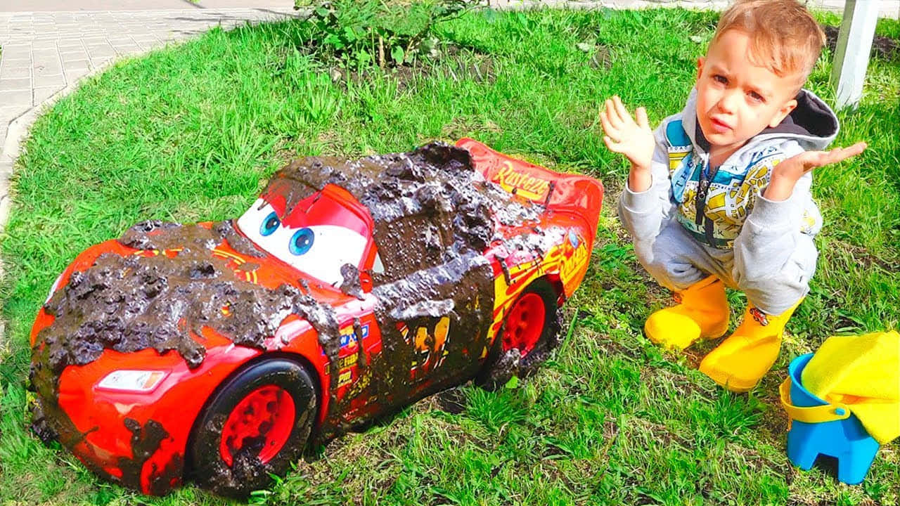 toys 桌布 壁紙 — 一個穿著黃色雨靴的幼兒蹲在草地上，旁邊是一輛沾滿泥巴的閃電麥坤玩具車。
