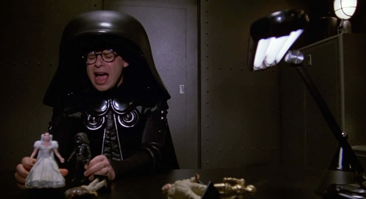 spaceballs 桌布 壁紙 — 一個穿著黑武士服裝的人坐在書桌前，在昏暗的房間裡，明亮的燈光下拿著星戰動作公仔。