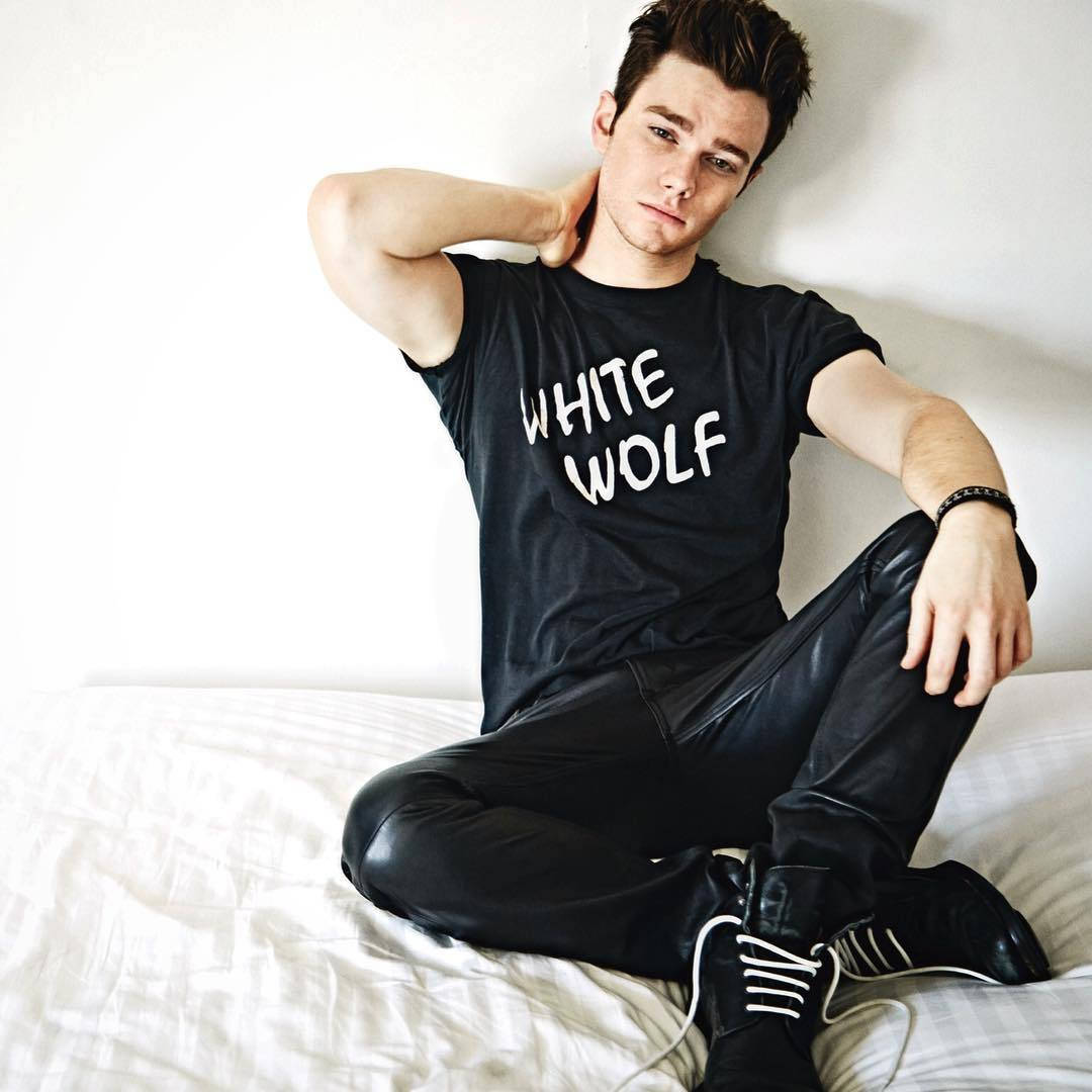 Chris Colfer 桌布 壁紙 — 一個穿著黑色「WHITE WOLF」T恤和皮褲的年輕男子，一手放在頸後，隨意地坐在白色床上。