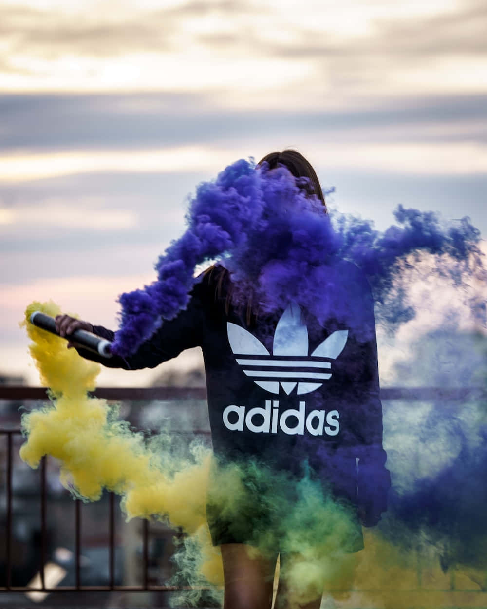 愛迪達 桌布 壁紙 — 一個穿著黑色Adidas連帽衫的人站在屋頂上，在柔和的暮色天空中釋放出充滿活力的紫色和黃色煙霧。