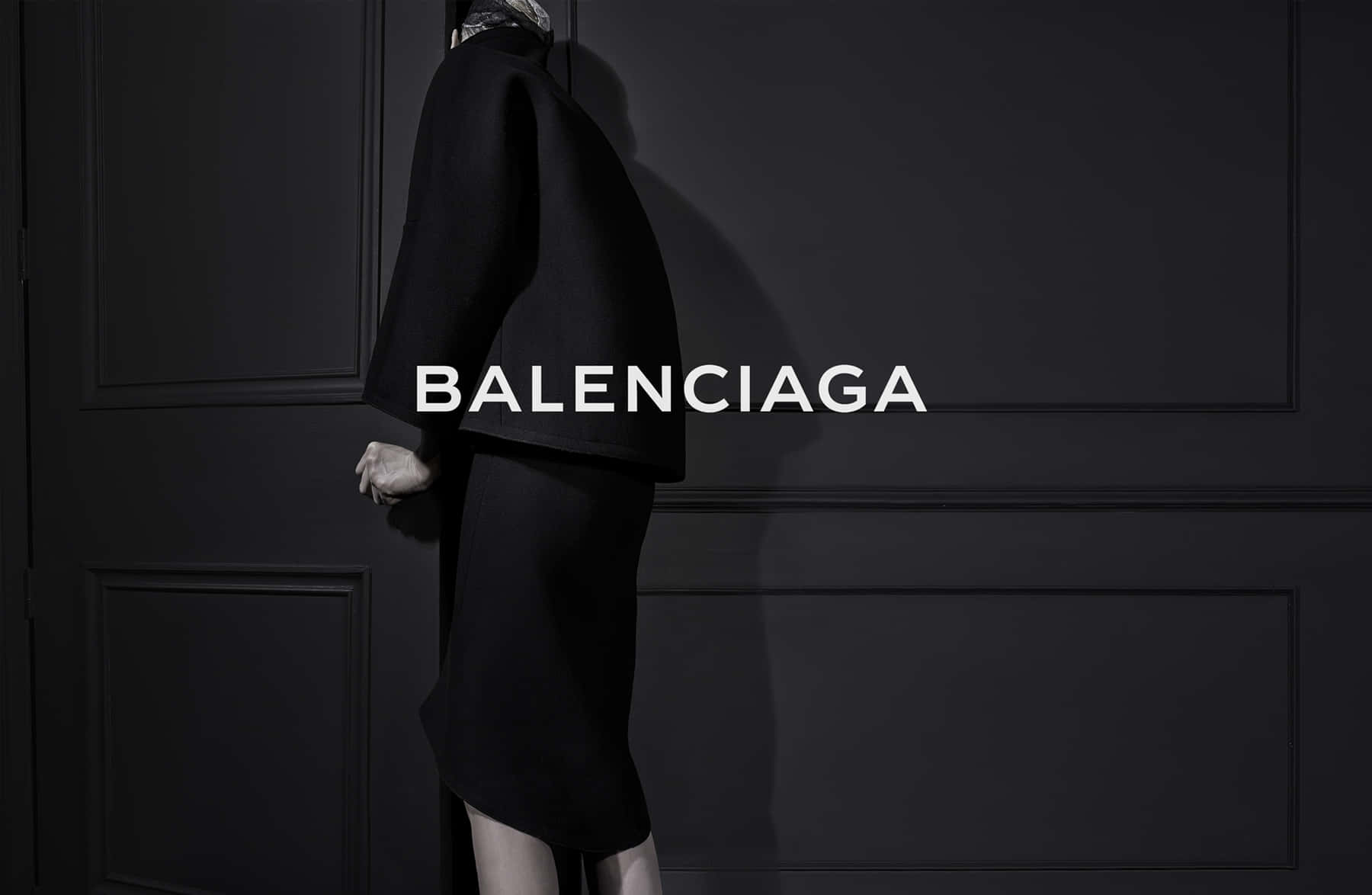 巴黎世家 桌布 壁紙 — 一個穿著黑色Balenciaga服裝的人側面站立在深色鑲板牆前，品牌名稱顯眼地展示。