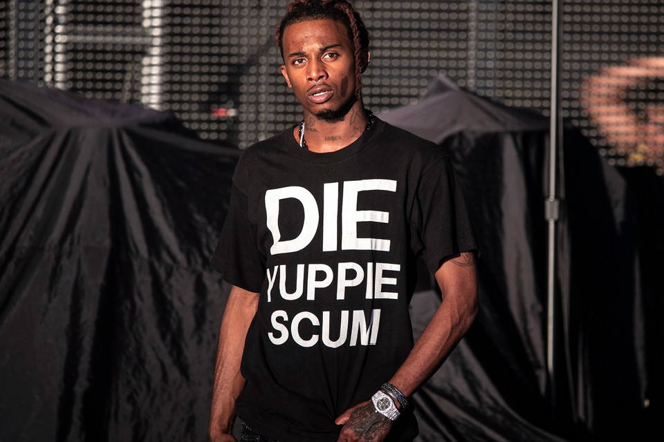 Carti 桌布 壁紙 — 一個穿著黑色T恤、上面有粗體白色文字的男人，自信地站在深色背景前，背景有垂墜布料和舞台燈光。