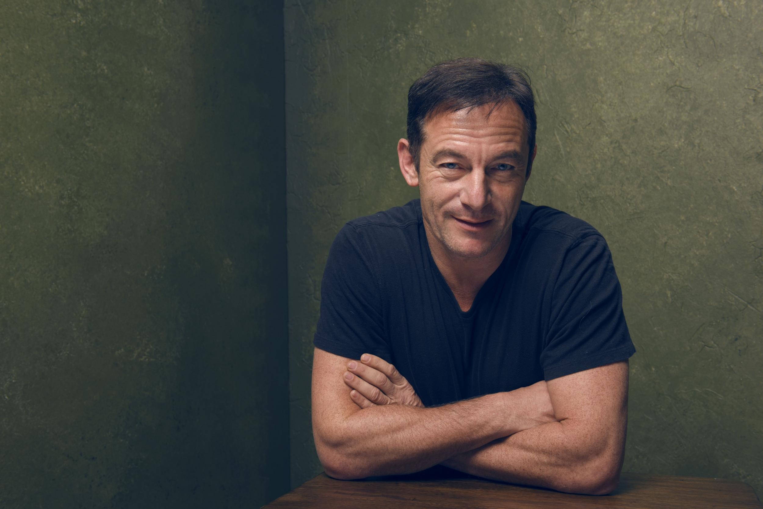actor 桌布 壁紙 — 一個穿著黑色T恤的男人，雙臂交叉，靠在一張木桌上，背景是帶有紋理的橄欖綠色牆壁。