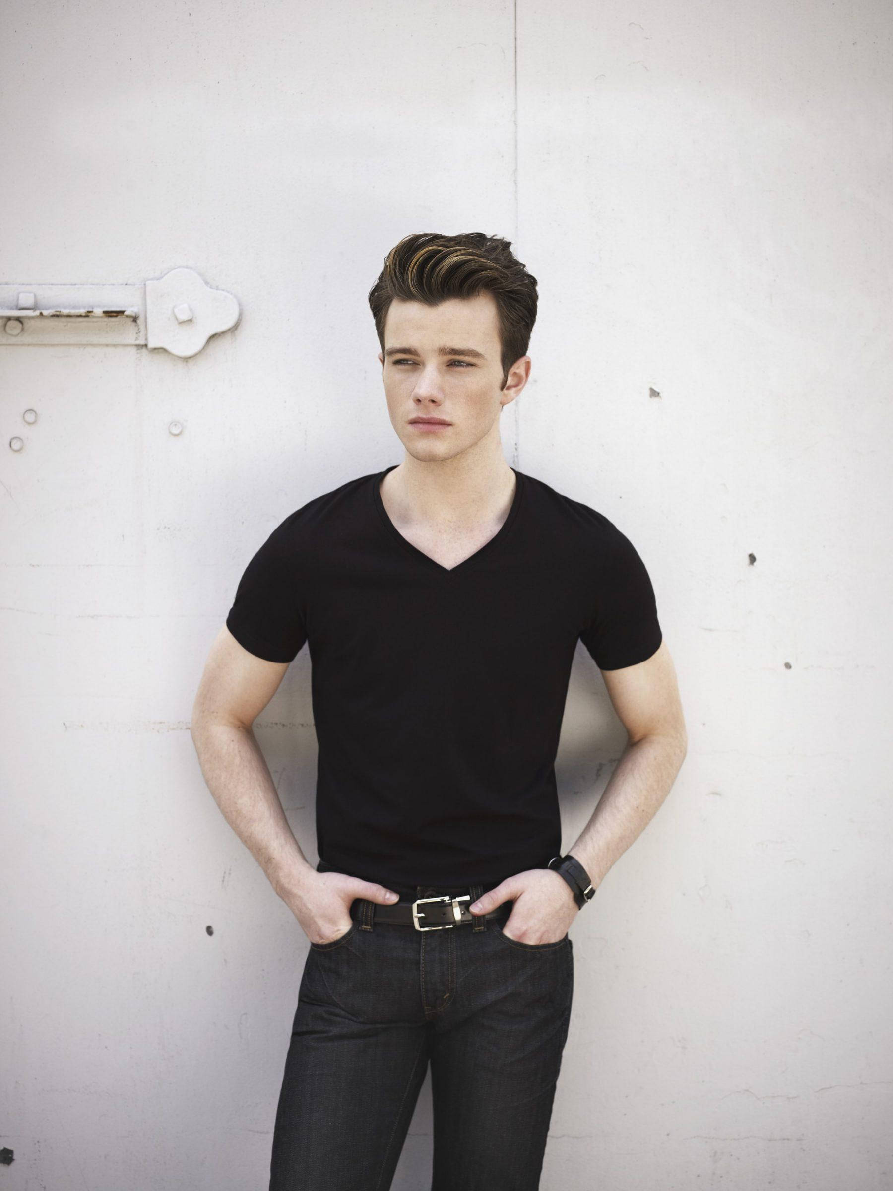 Chris Colfer 桌布 壁紙 — 一個穿著黑色V領襯衫和牛仔褲的年輕男子，雙手插袋，隨意地靠在一面風化的白牆上。