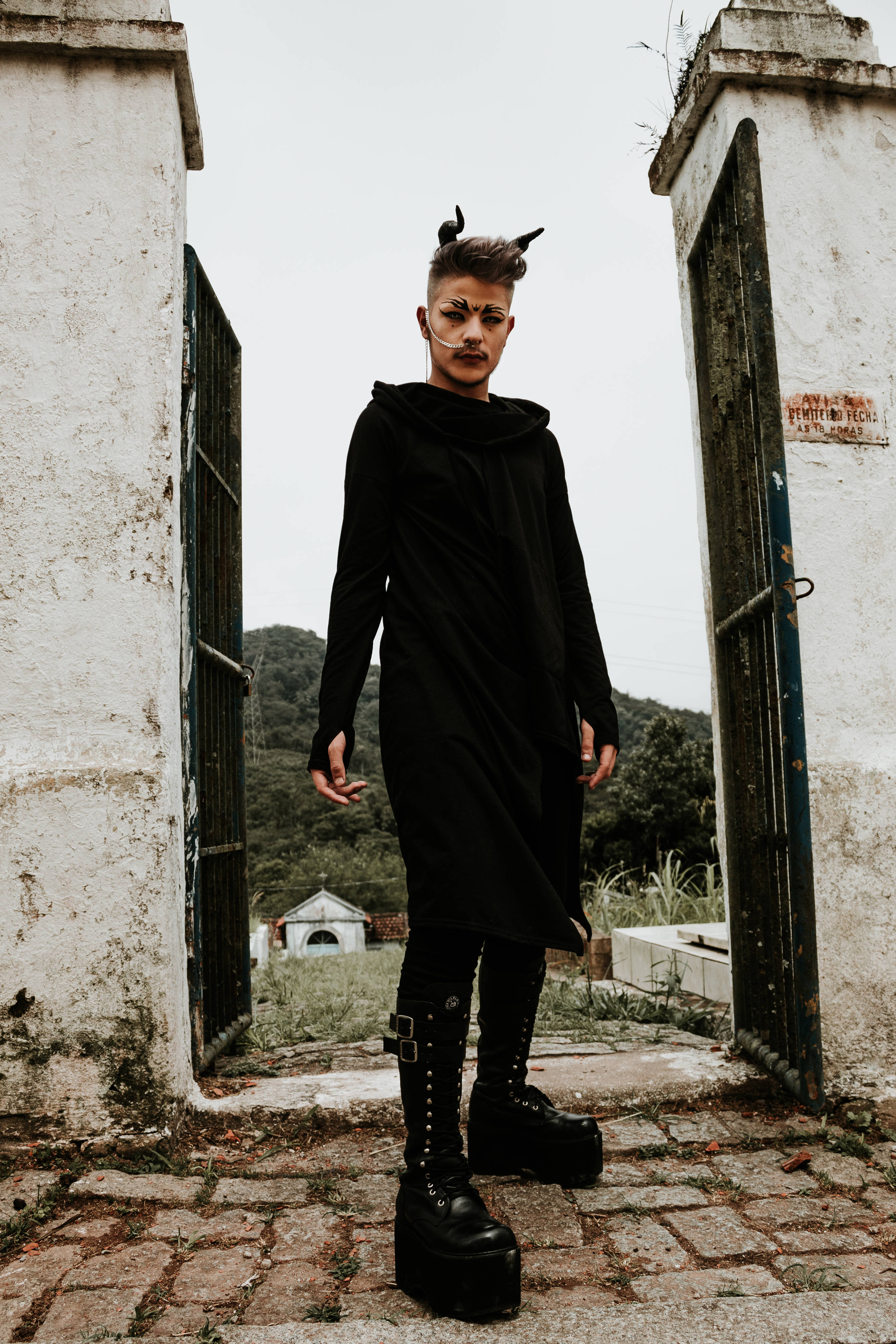 Goth 桌布 壁紙 — 一個穿著黑色哥德式服裝、帶有角飾的人站在一個風化的石門中，周圍環繞著茂密的植被。