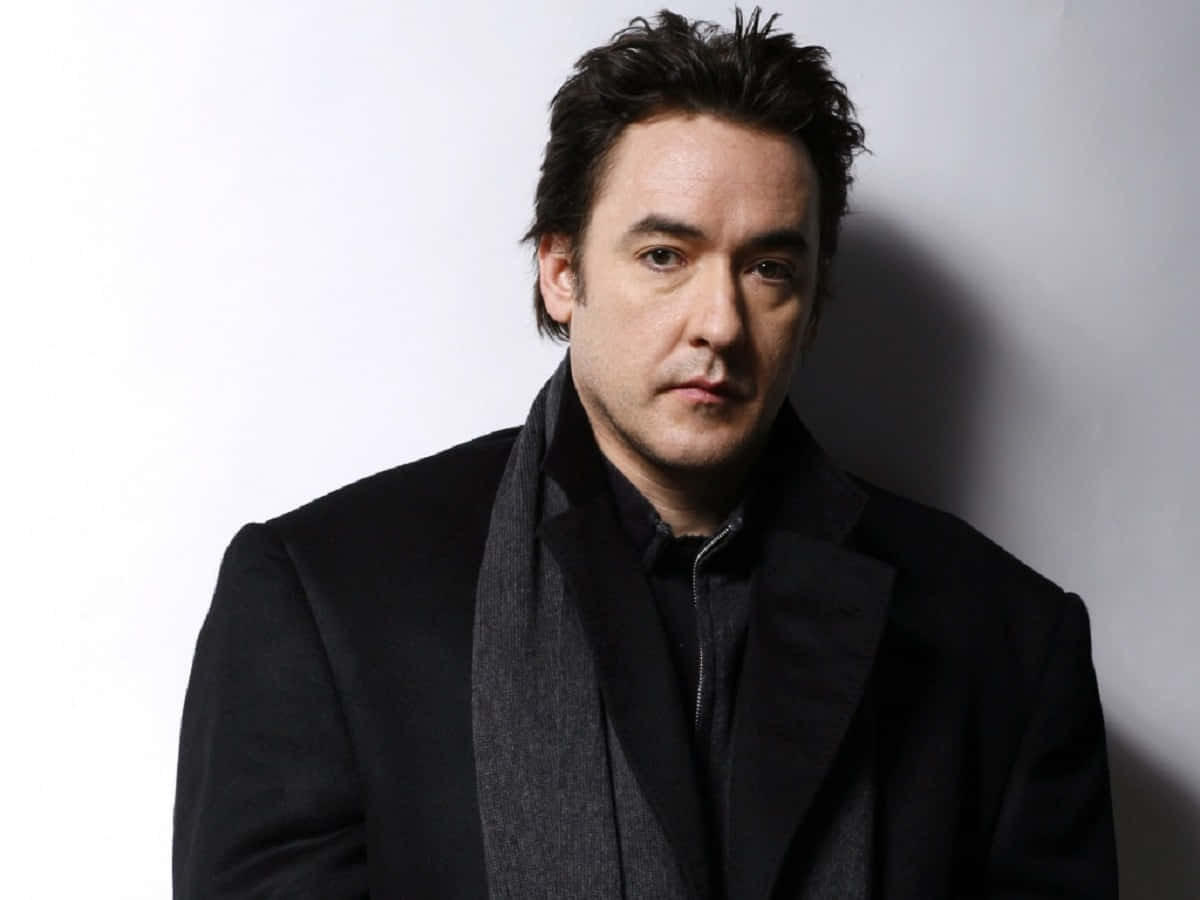 John Cusack 桌布 壁紙 — 一個穿著黑色外套和圍巾的男人站在純白色牆壁前，戲劇性的燈光投射出陰影。
