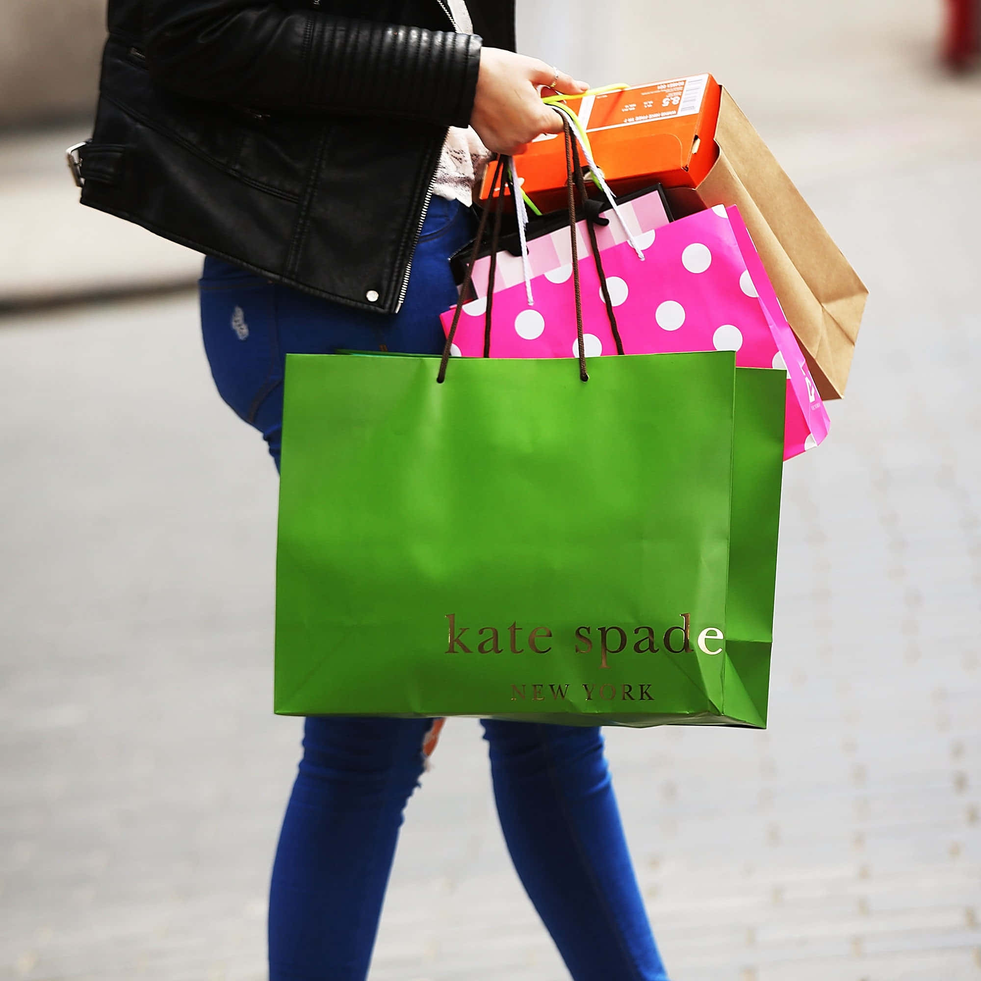 Kate Spade 桌布 壁紙 — 一個穿著黑色外套和藍色牛仔褲的人，在鋪砌的人行道上拿著多個色彩繽紛的購物袋，包括一個顯眼的綠色Kate Spade手提袋。