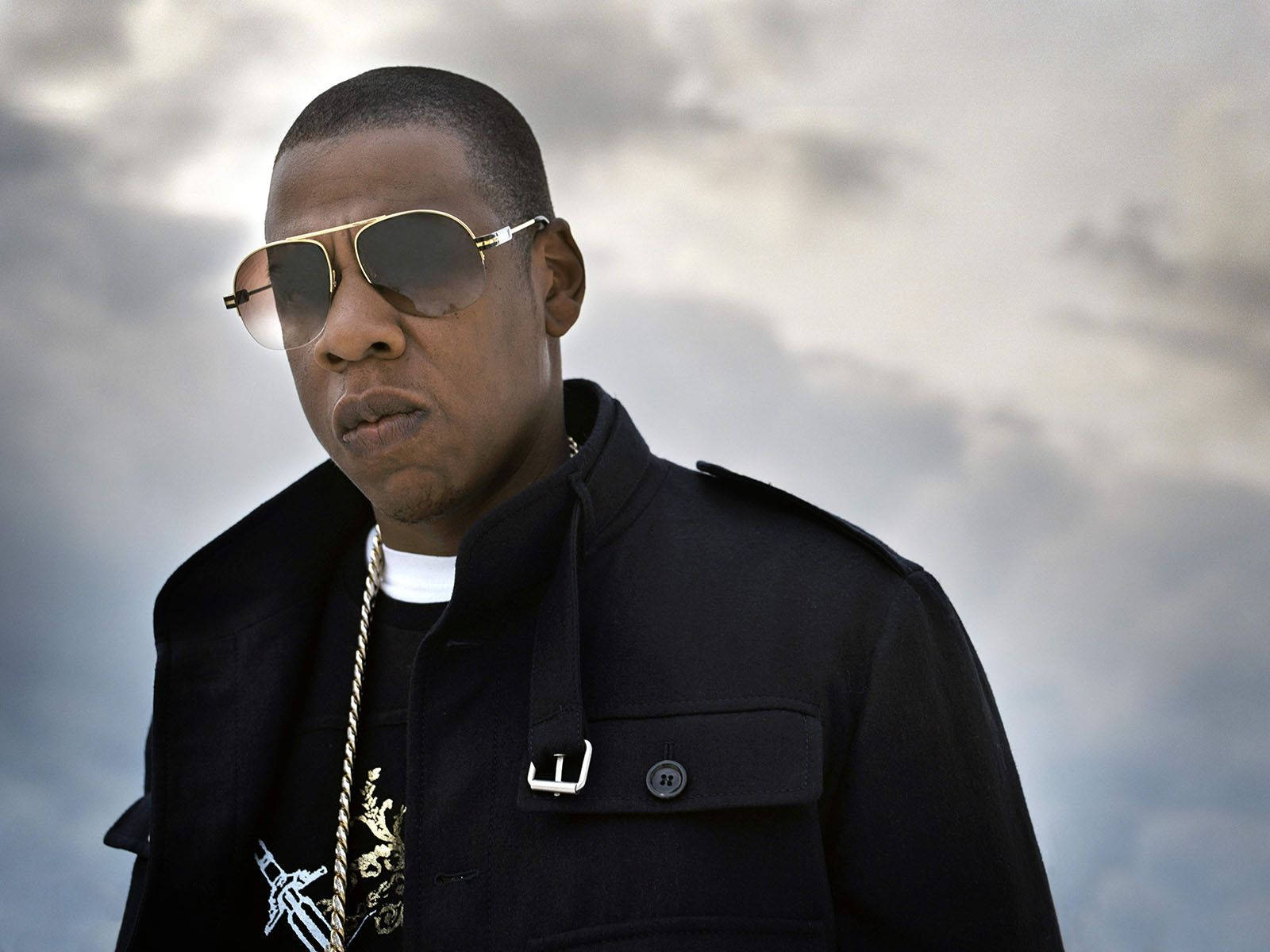 Jay-z 桌布 壁紙 — 一個穿著黑色外套和金色邊框太陽眼鏡的男人，在陰天的天空下帶著嚴肅的表情站立。