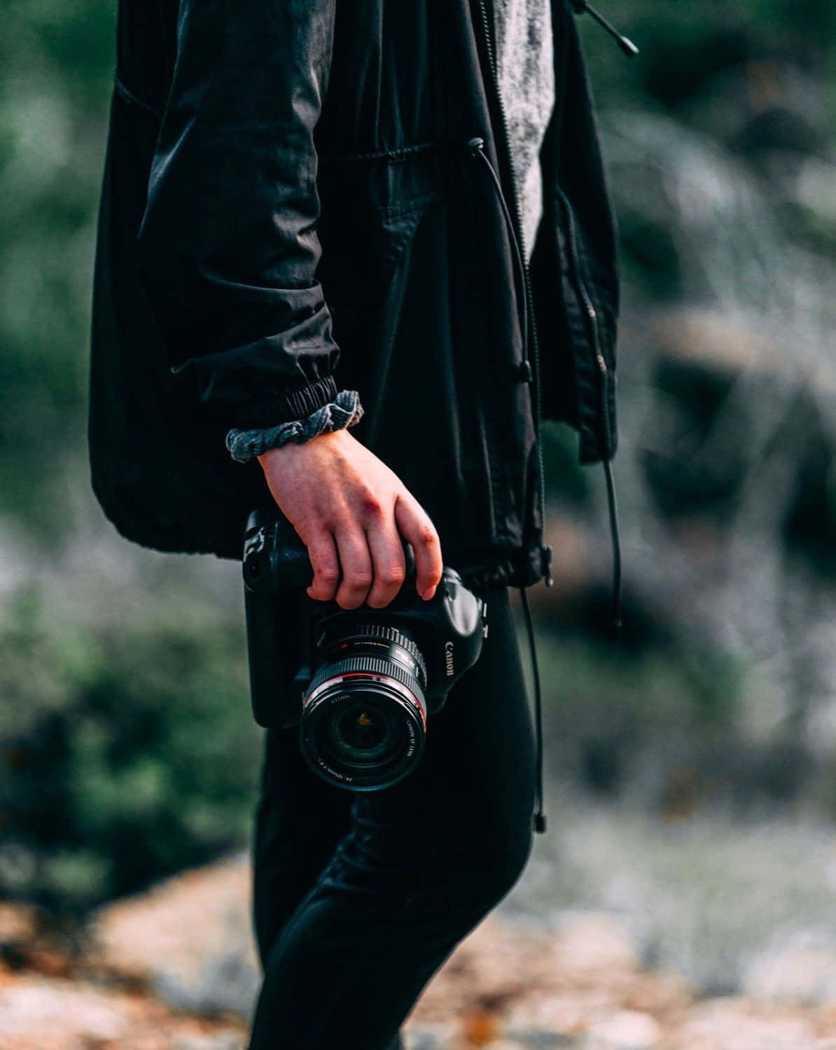 Holding Camera 桌布 壁紙 — 一個穿著黑色外套的人，在模糊的自然風景中行走時拿著相機。
